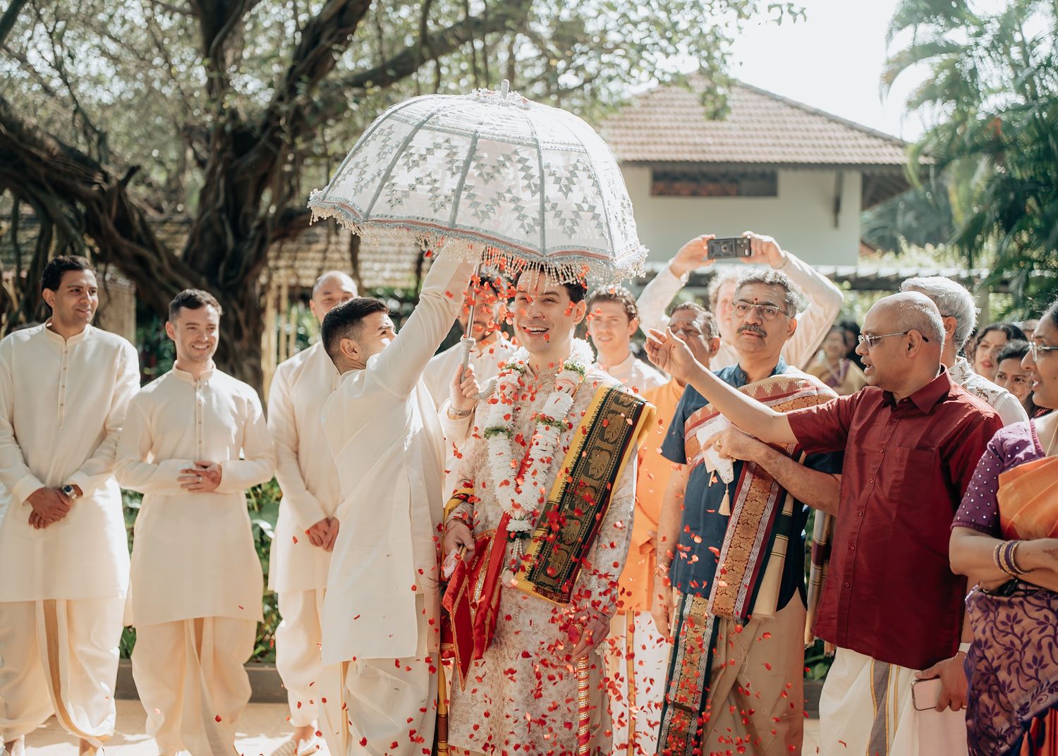 Groom in traditional Indian wedding attire walks under umbrella as guests shower flower petals outdoors.
