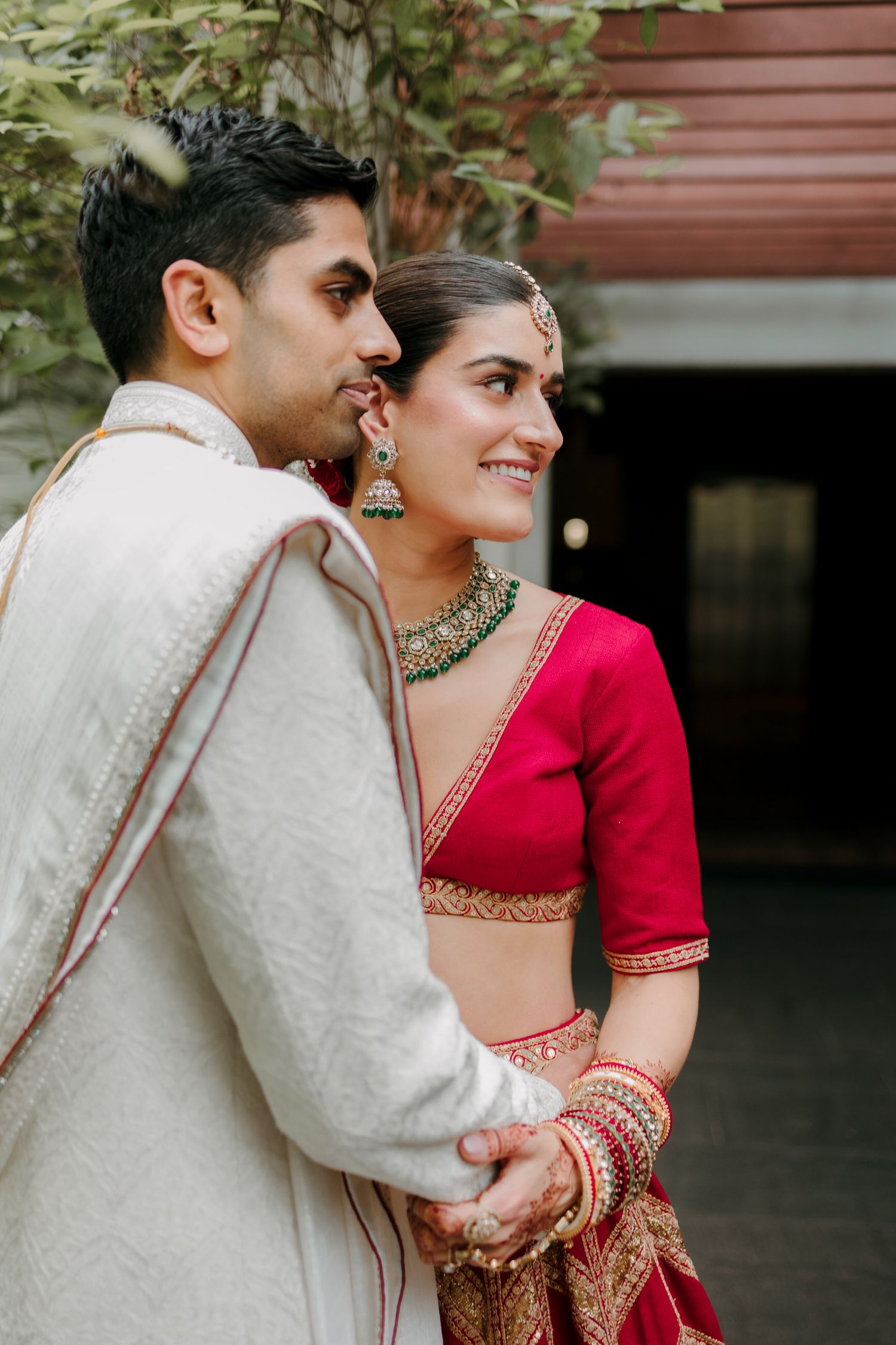 Indian bride in red lehenga and groom in white sherwani holding hands and smiling at their traditional wedding.