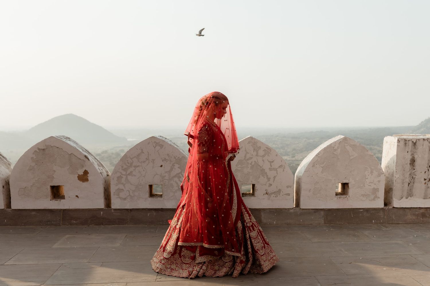 Silhouette of bride in red lehenga against white architecture and mountain backdrop.