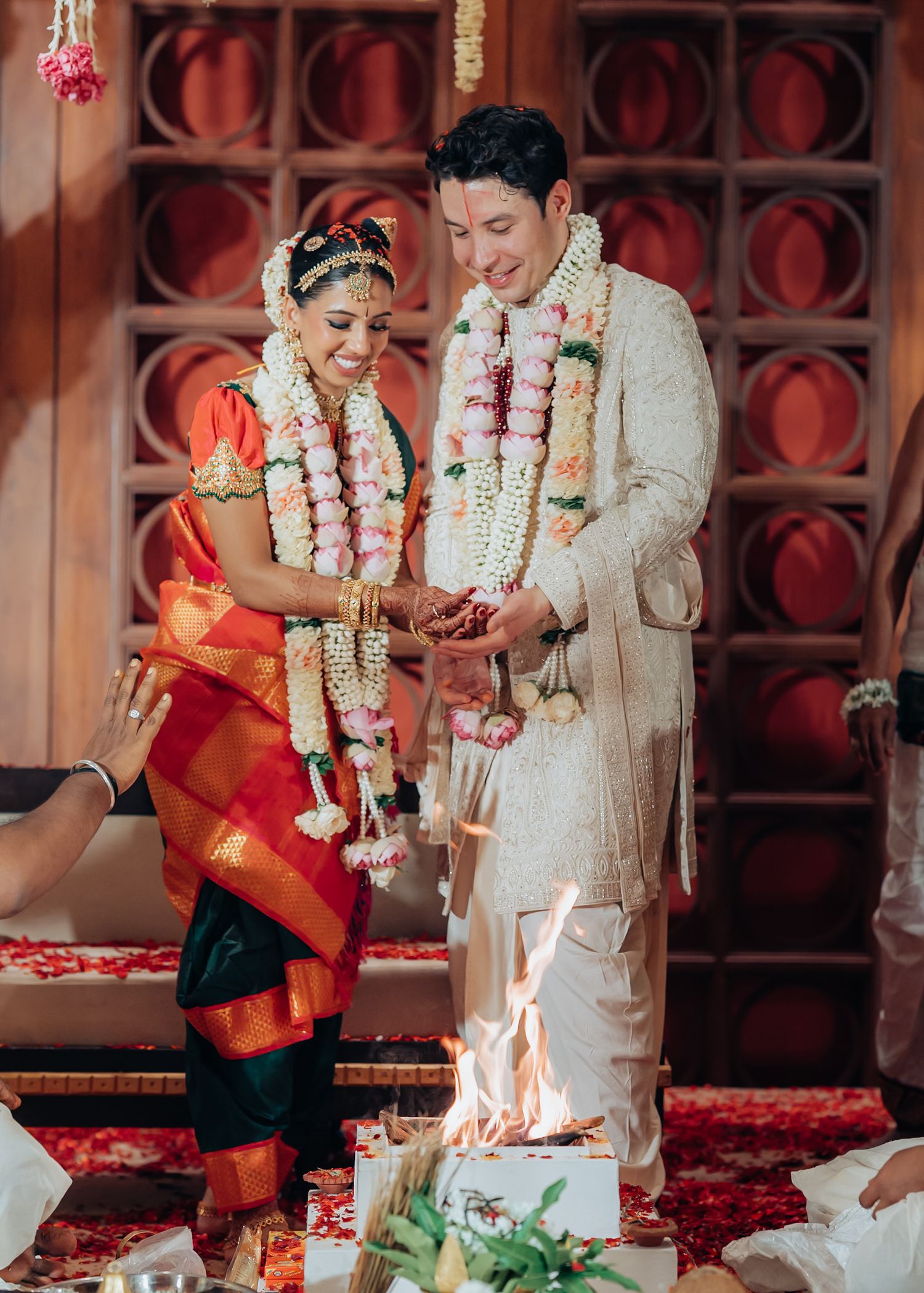 Wedding ritual being performed with flower garlands against an ornate red backdrop.