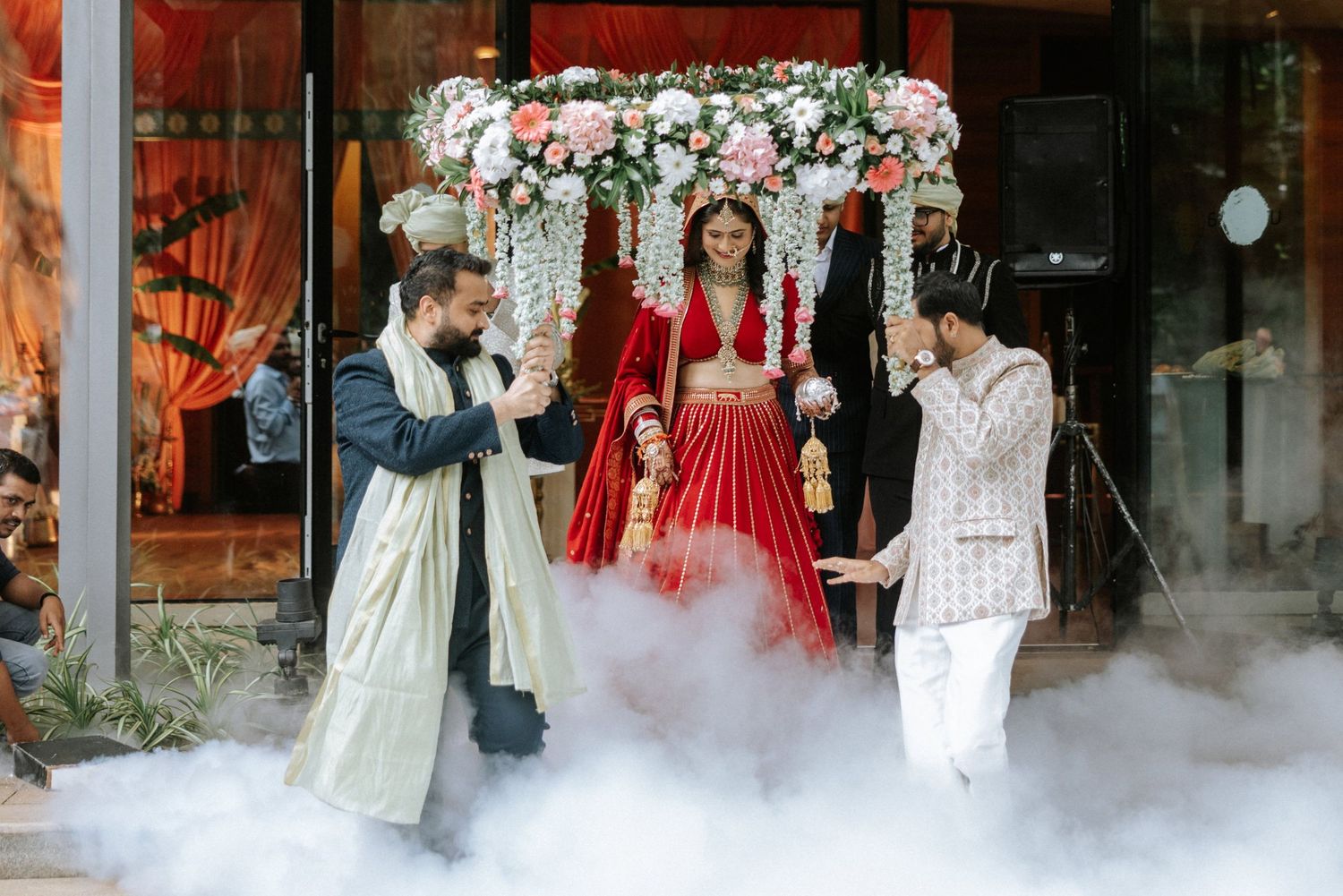 Bride in red lehenga carried under floral canopy with smoke effects at Indian wedding ceremony.