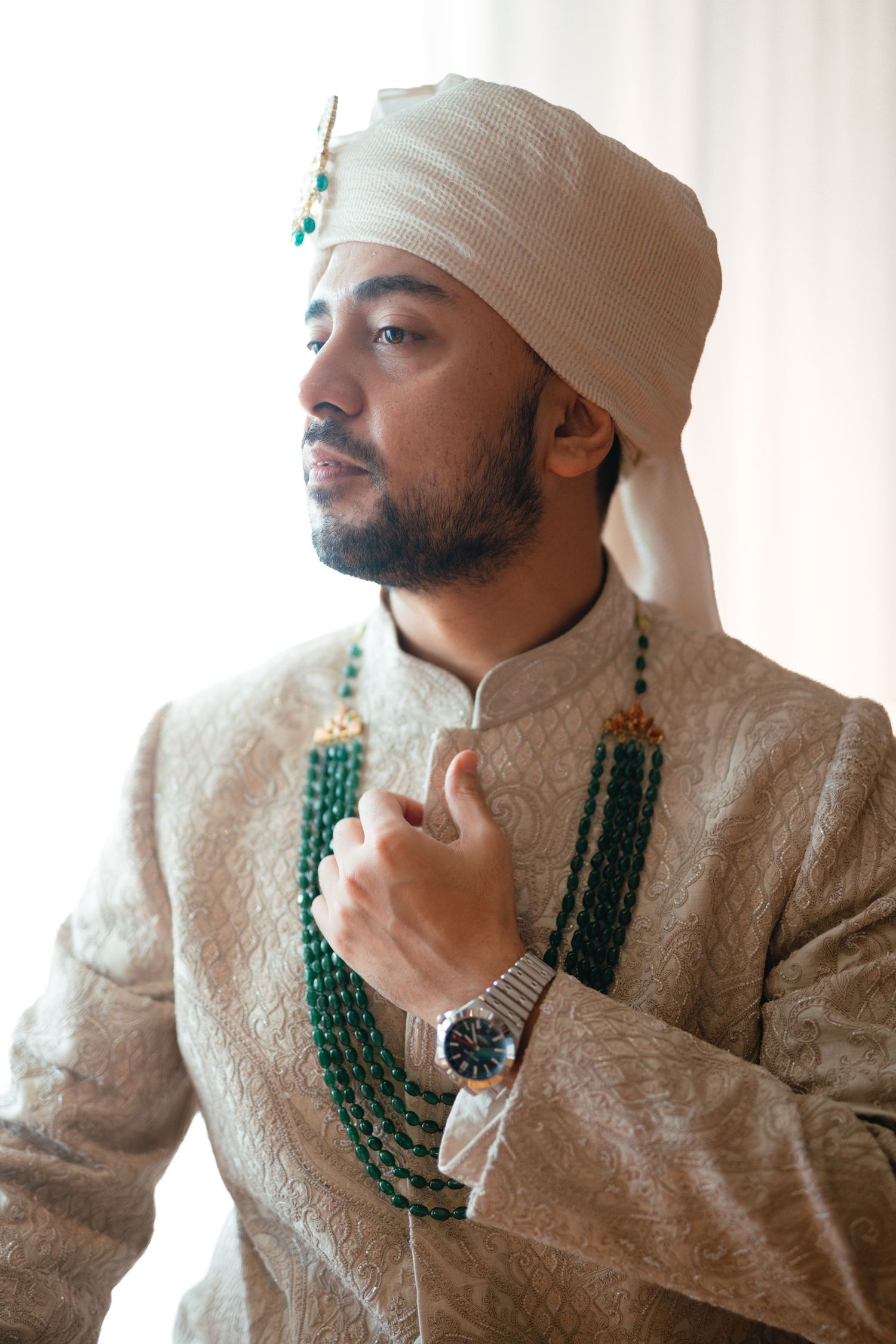 Indian groom in embroidered sherwani, white turban, emerald beaded necklace, and luxury watch at wedding.