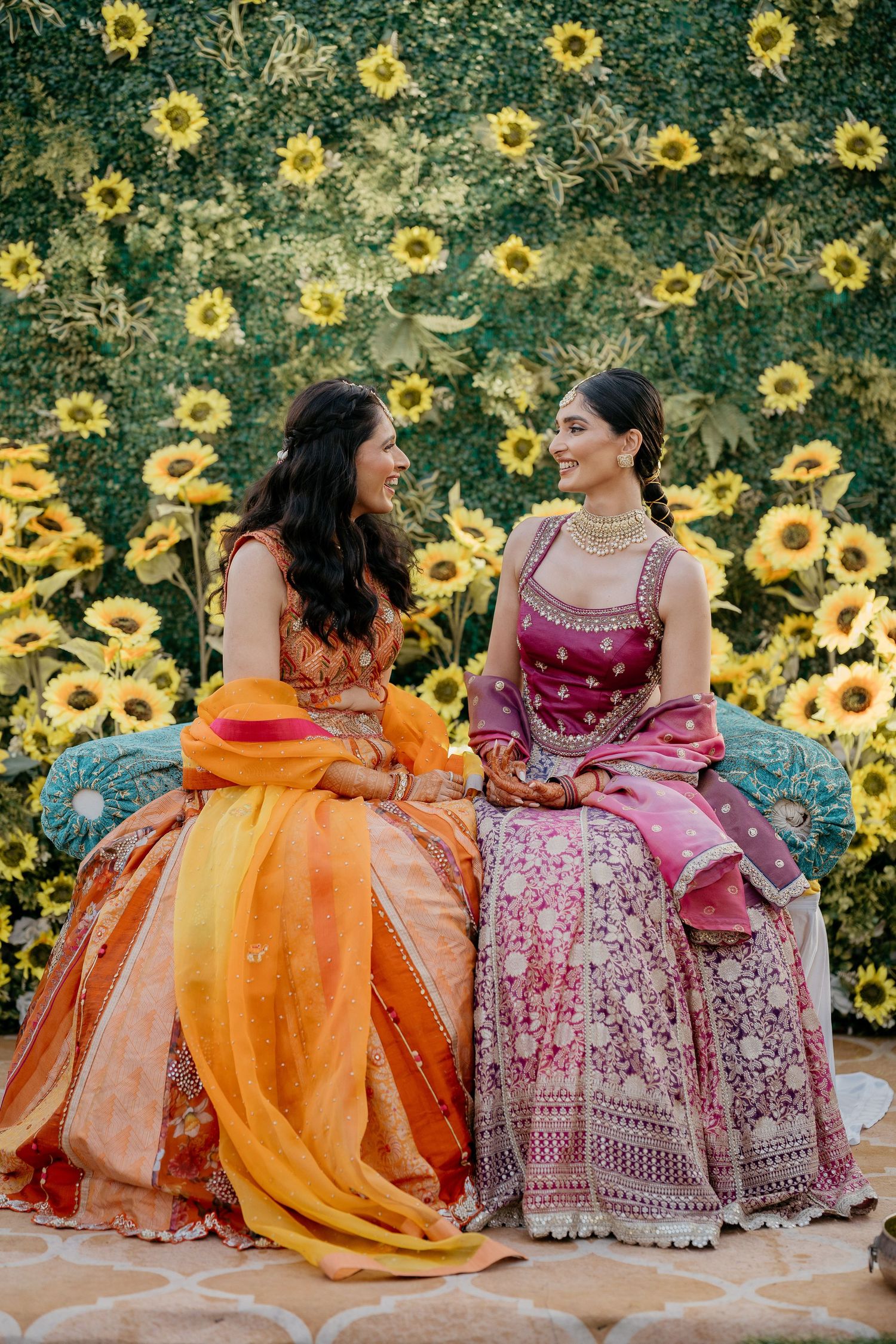 Bride and her sister seated together in traditional Indian lehengas against a vibrant sunflower backdrop at Taj Lands End Mumbai wedding