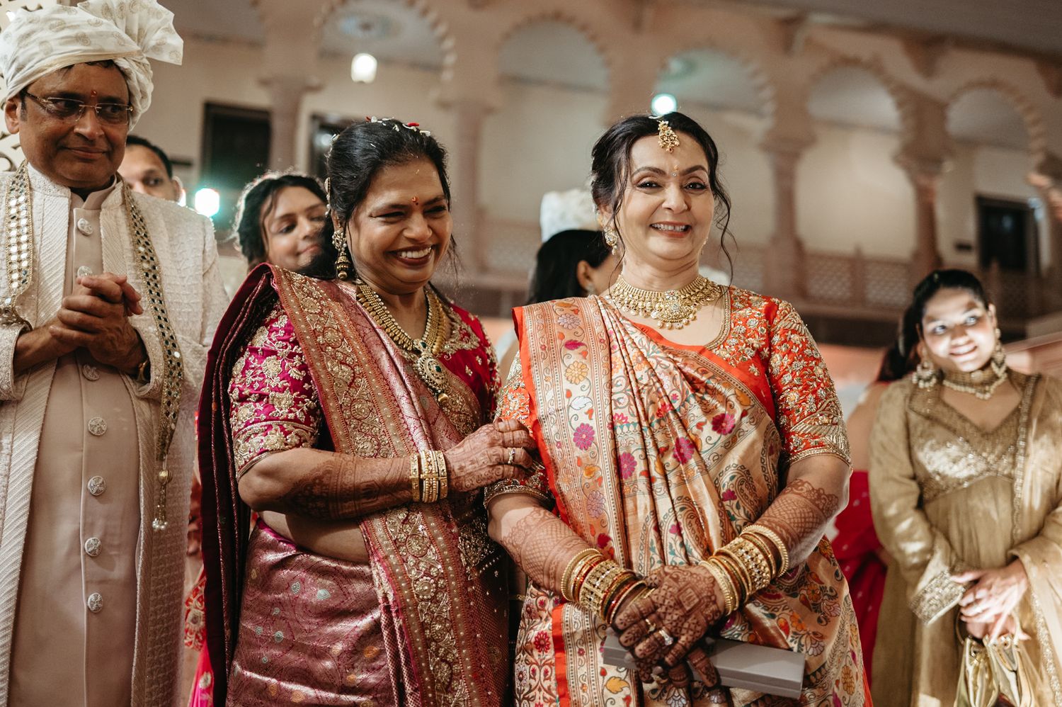 Bride in red and gold saree smiles with family during traditional Indian wedding ceremony.