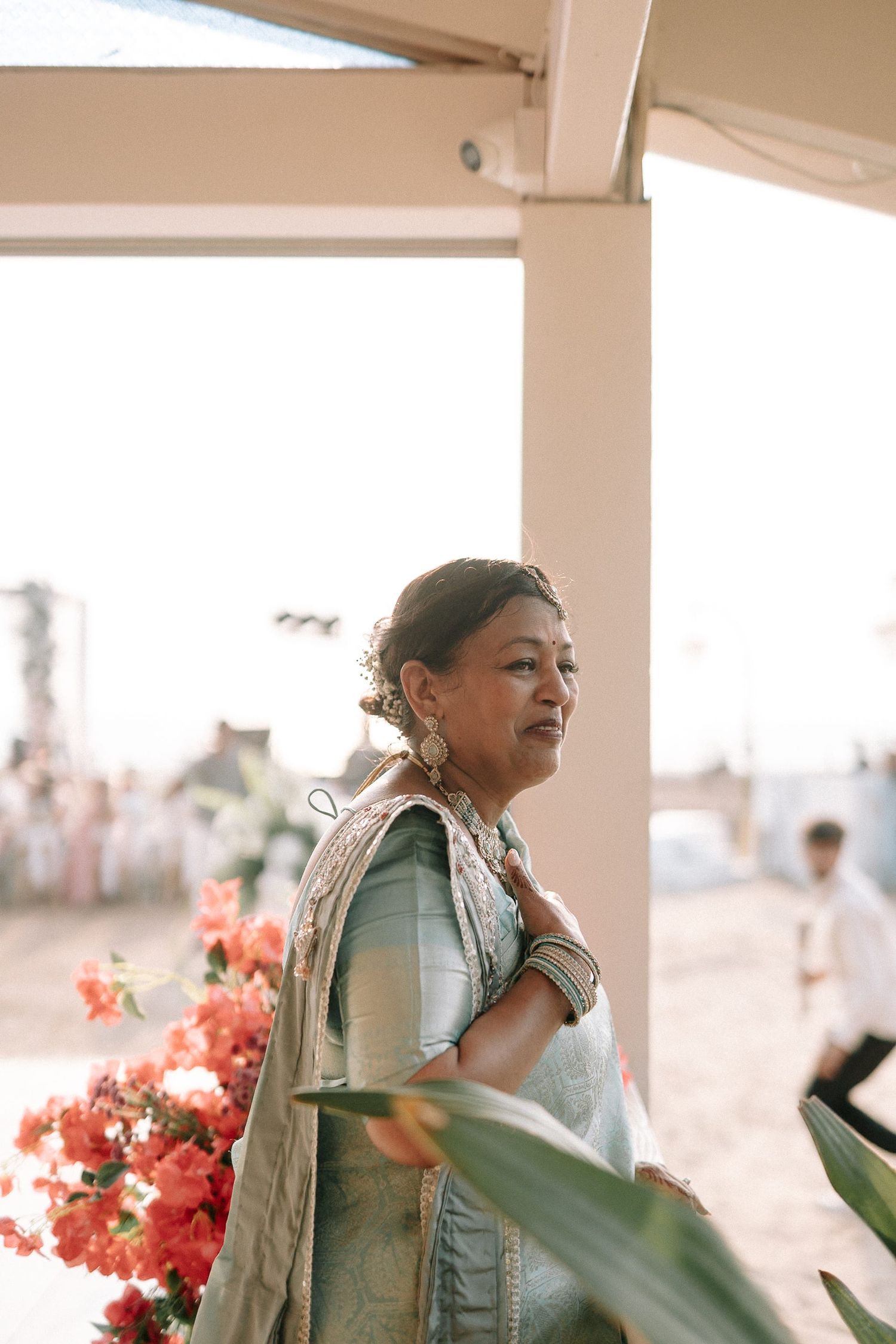 Elegant Indian woman in light blue saree with gold embroidery stands near red flowers at wedding.