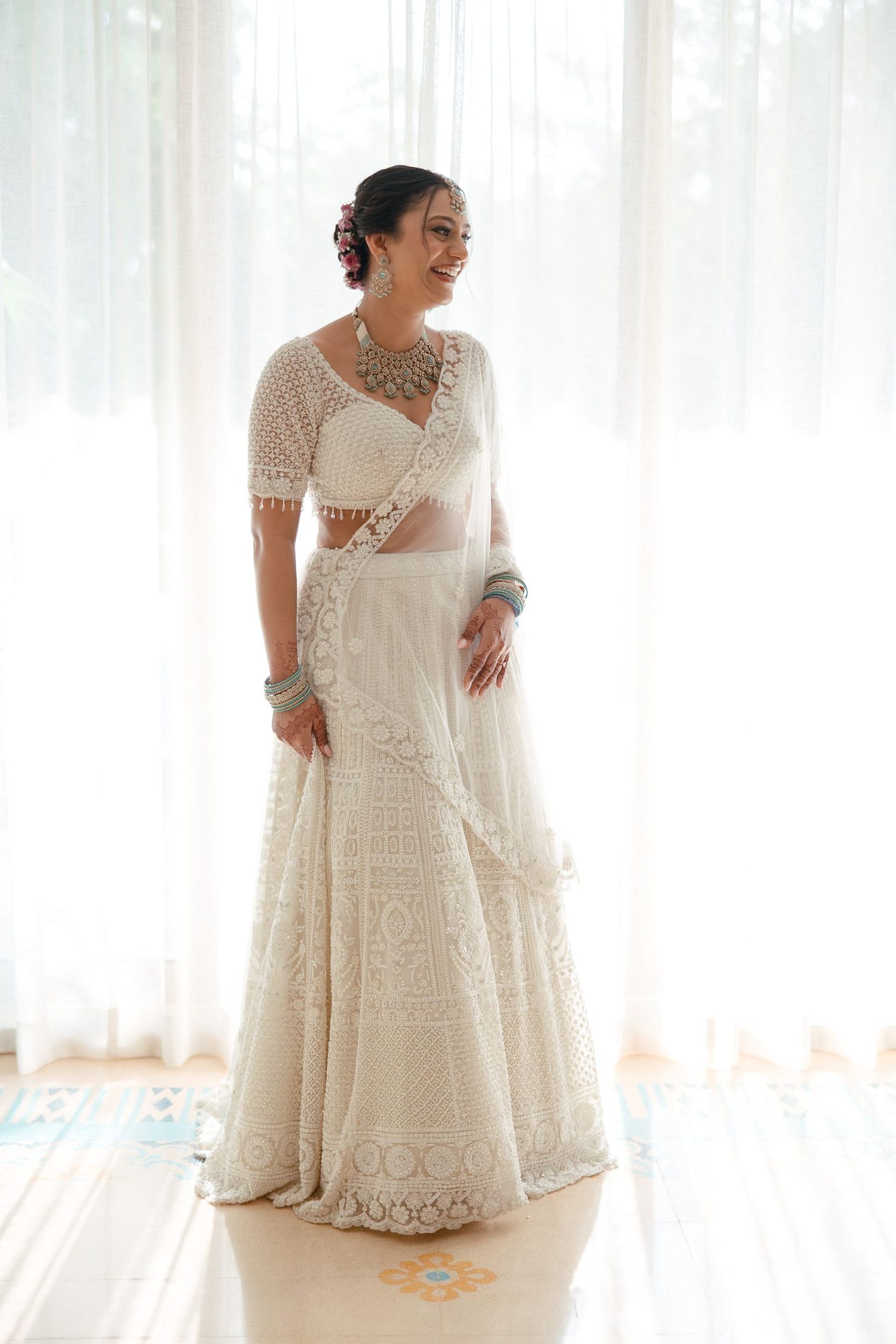 Bride in white embroidered lehenga choli with sheer dupatta, turquoise jewelry, posing by sheer curtains.