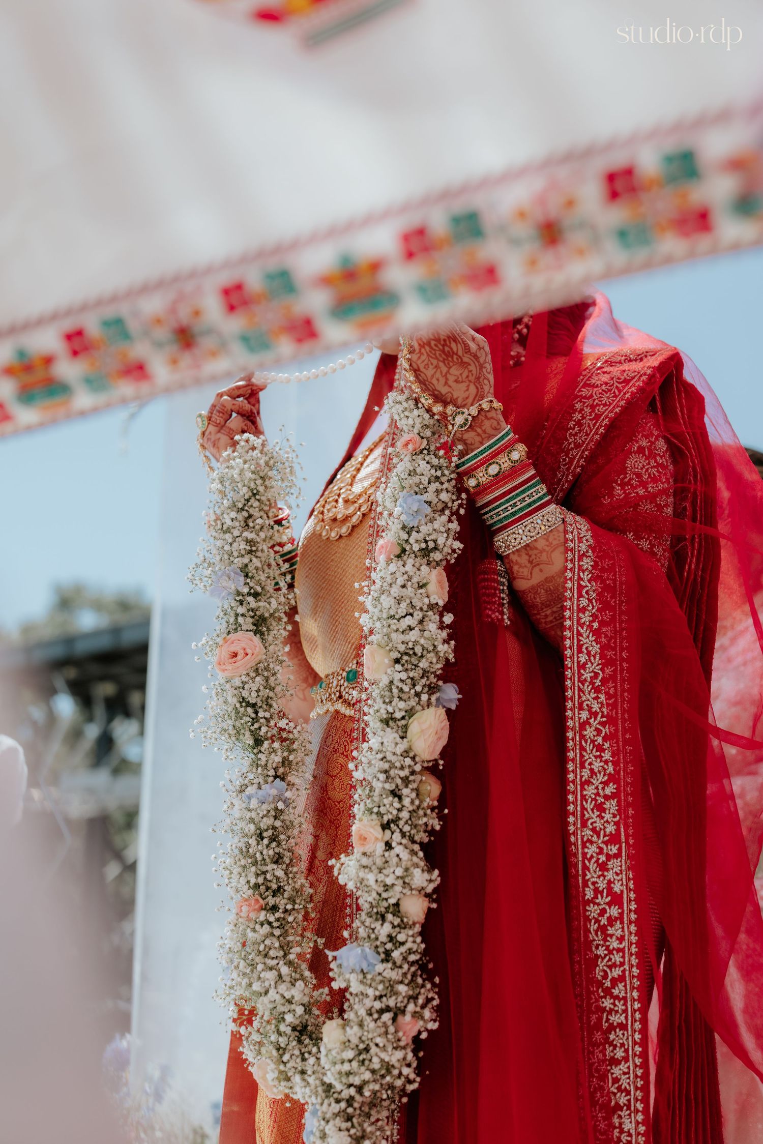 Bride in red lehenga holding floral garland of white baby's breath and roses at Indian wedding ceremony.