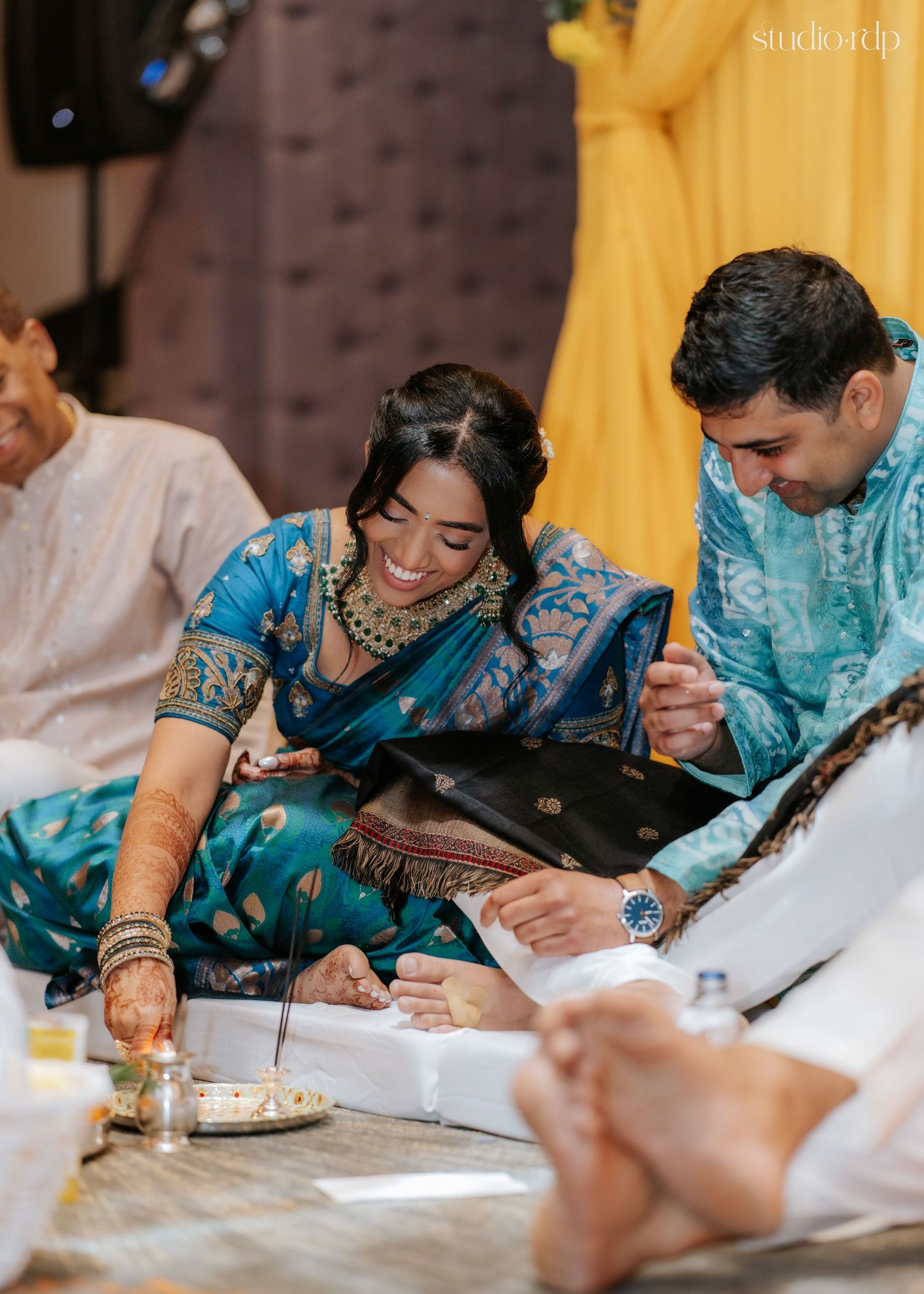 Couple in teal Indian attire smiling during traditional wedding ceremony ritual, woman in silk saree with gold jewelry.
