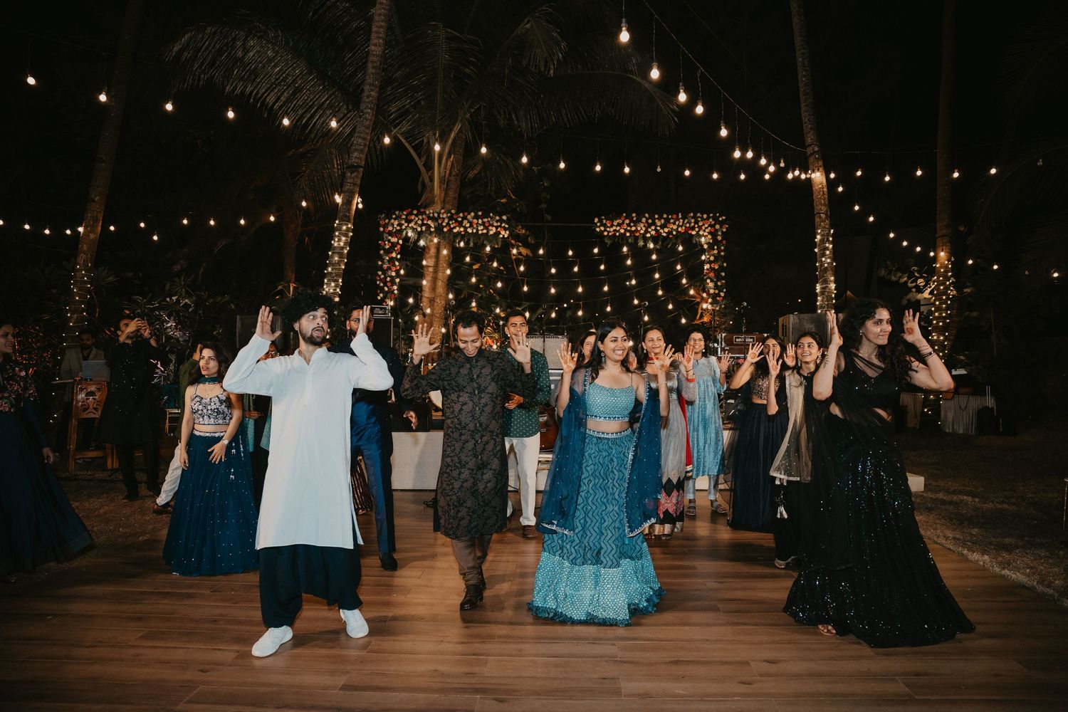 Guests dancing at an outdoor Indian wedding reception with string lights and palm trees at night.