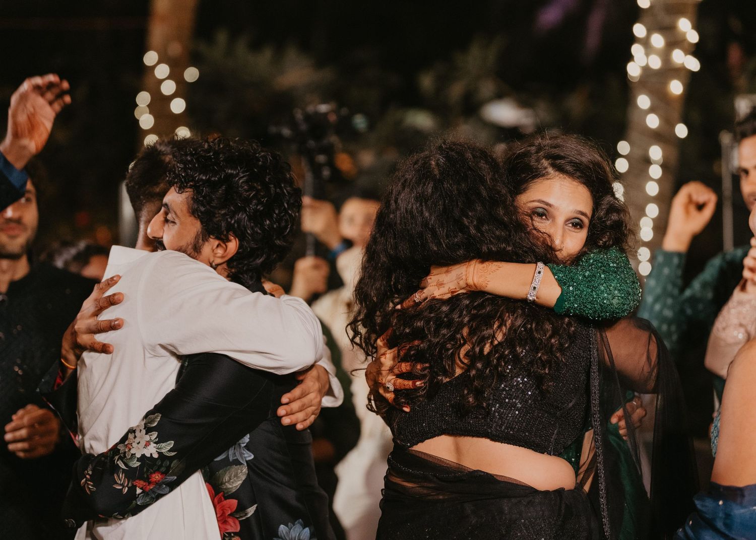 Guests embracing and dancing at an outdoor evening celebration with festive string lights in the background.