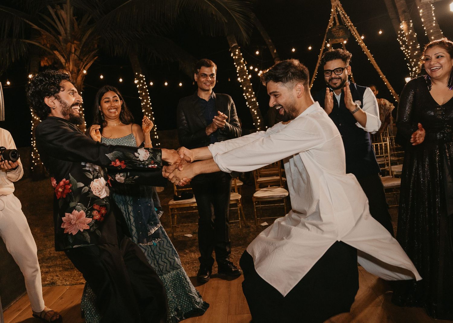 Two men dancing joyfully at an outdoor wedding reception surrounded by guests and string lights.