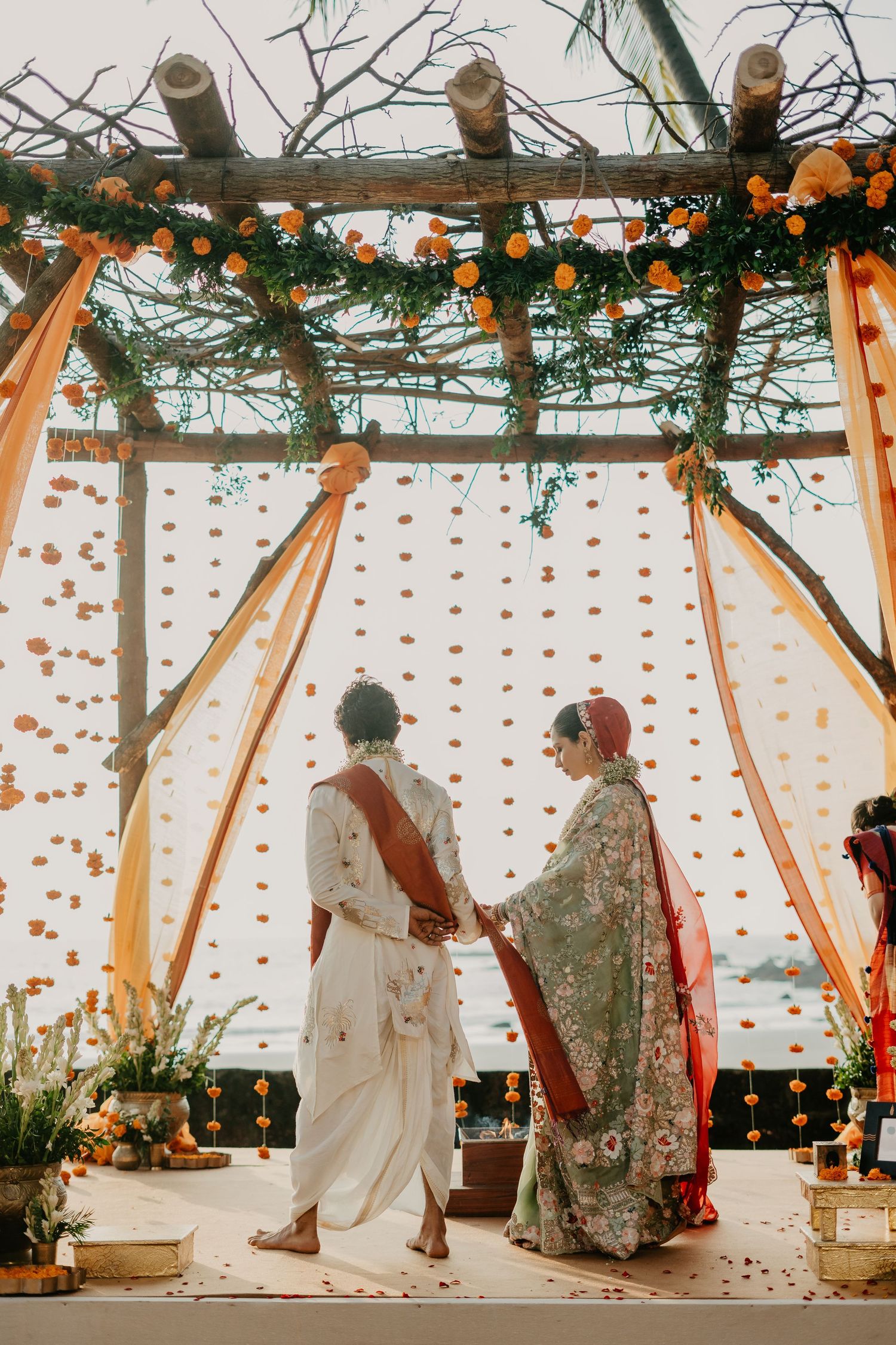 Couple holding hands during Indian wedding ceremony under floral mandap with marigold decorations.
