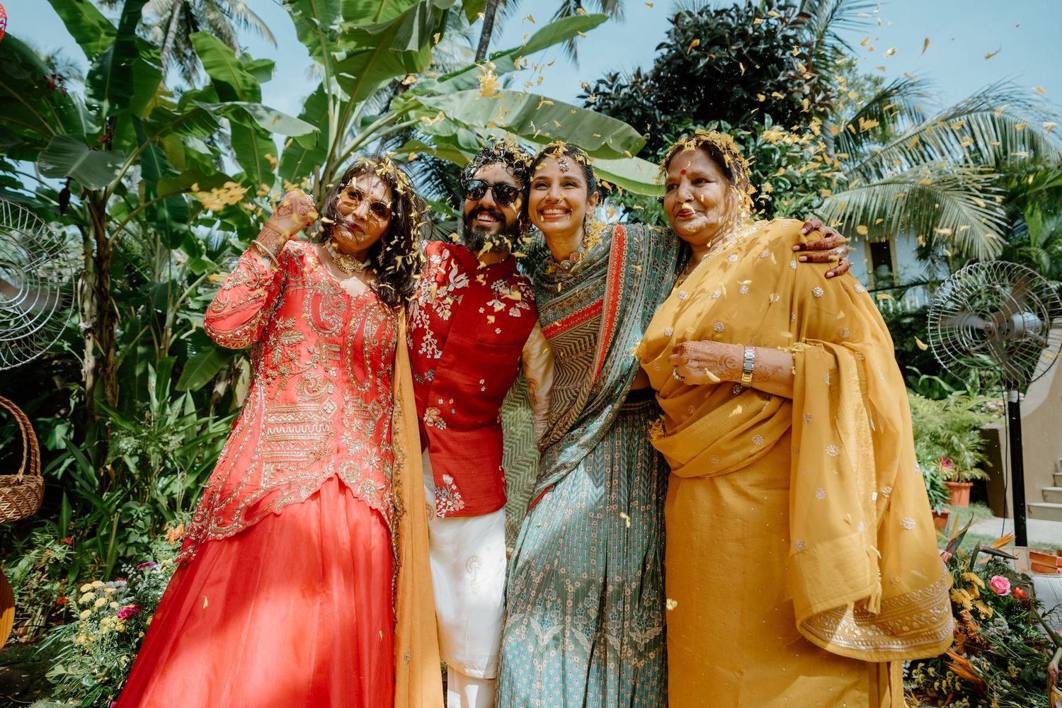 Four women in vibrant traditional Indian attire smile together outdoors amid lush tropical greenery.