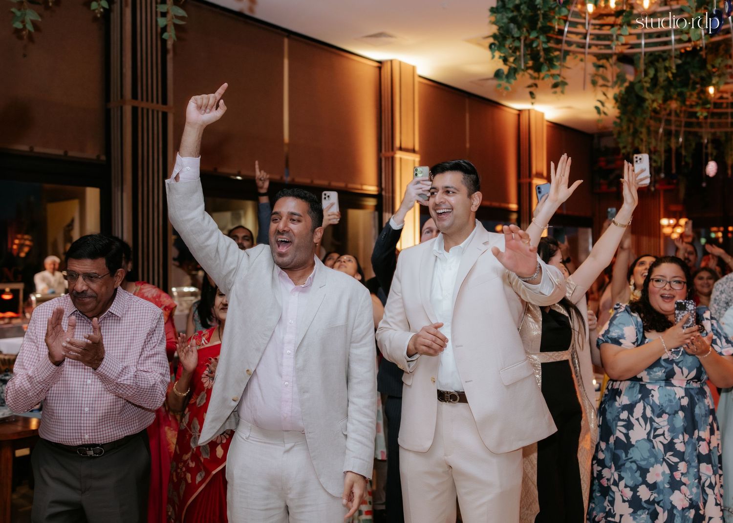 Two men in white suits celebrate with raised arms at a festive indoor wedding reception with guests cheering behind them.