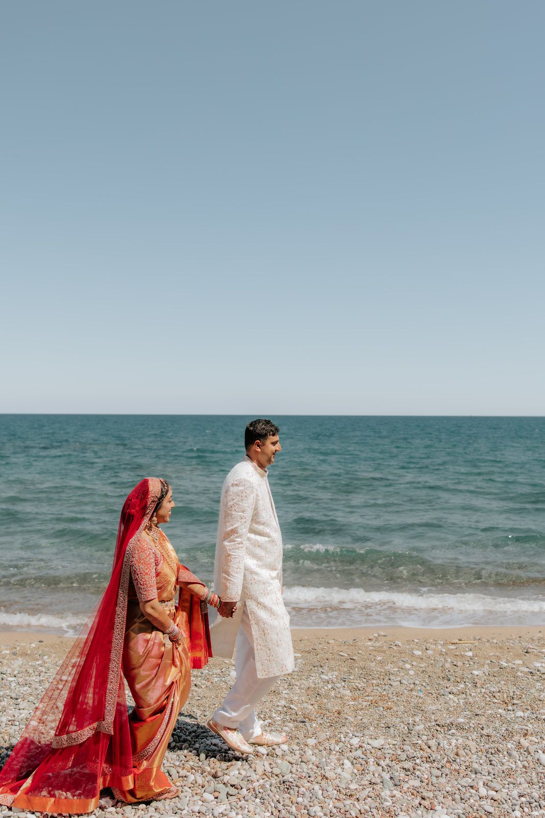 Indian couple in traditional wedding attire holding hands on a rocky beach with calm turquoise sea backdrop.