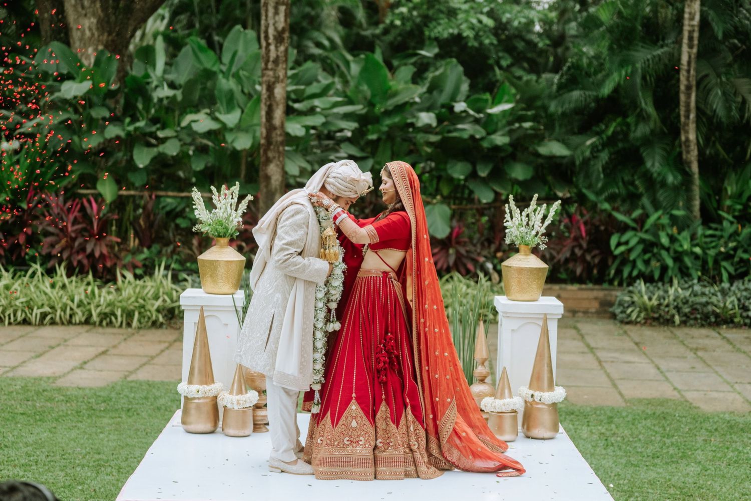 Indian bride in red lehenga and groom in white sherwani share a romantic moment during outdoor wedding ceremony.