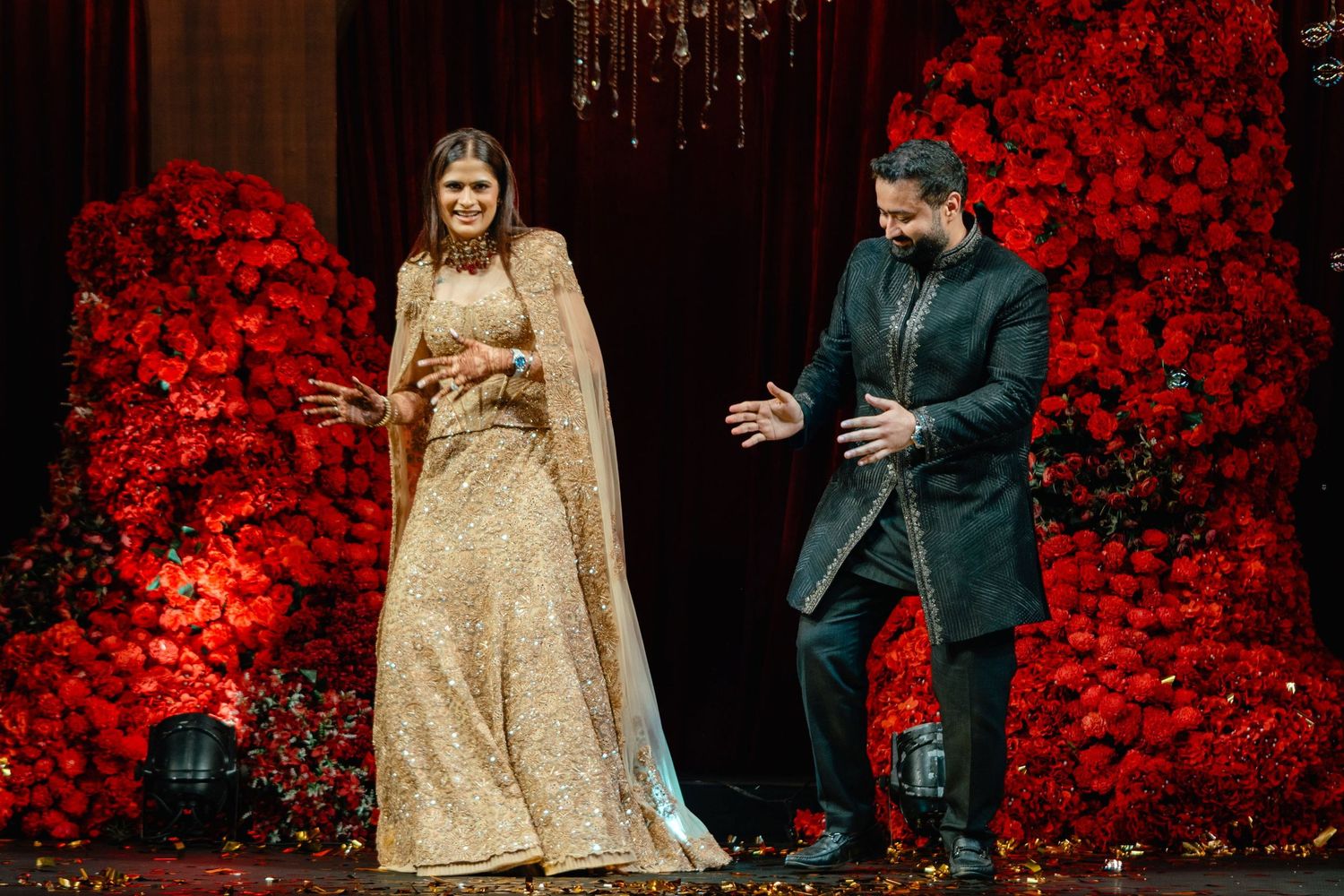 Bride in gold lehenga and groom in dark sherwani stand on stage surrounded by red floral arrangements.