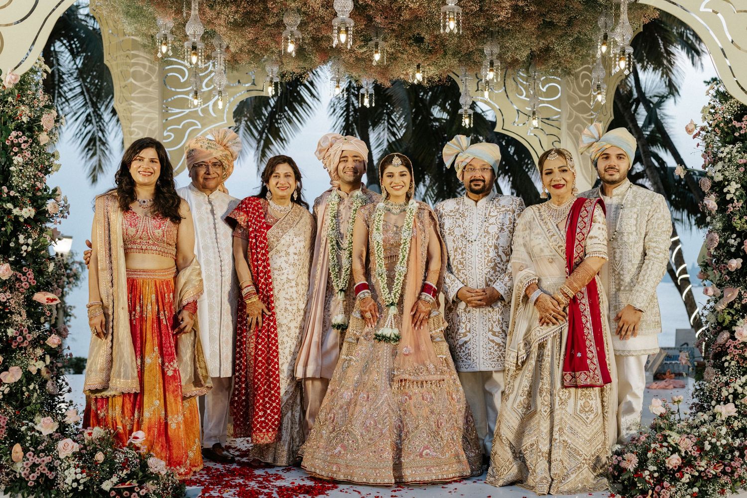 A wedding party in traditional Indian attire poses together under an ornate floral arch at an elegant ceremony.