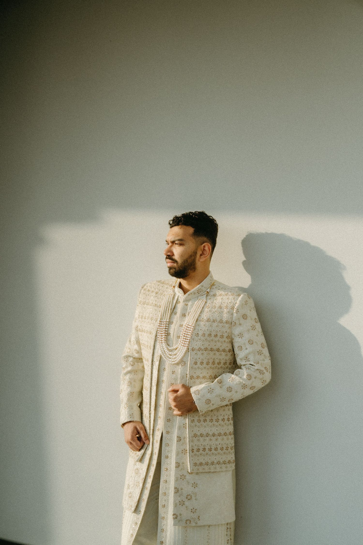 Person wearing an elegant cream-colored embroidered traditional Indian sherwani poses against a neutral wall.