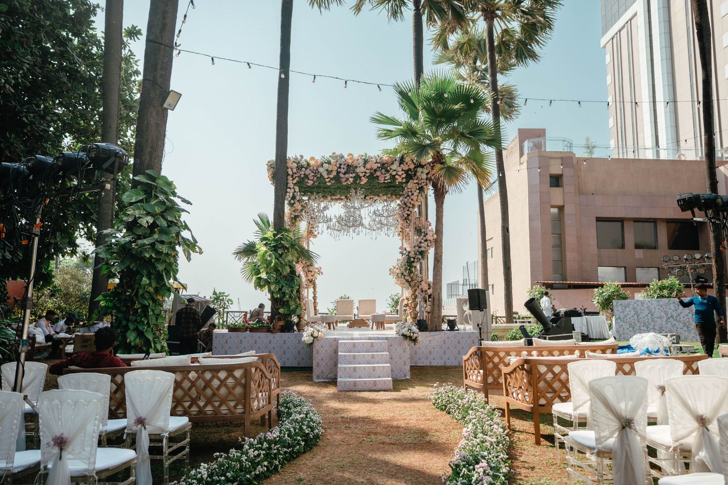 Rustic outdoor venue decorated with string lights, palm trees, and white event furniture creates a tropical paradise setting.