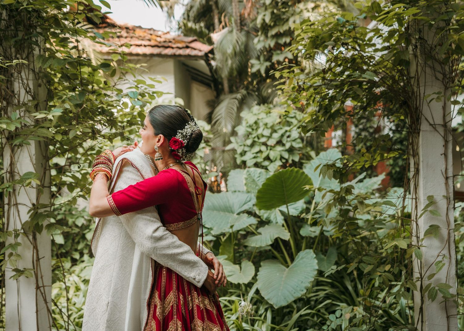 A romantic garden scene with a couple in elegant red and white attire sharing a tender moment among lush greenery.