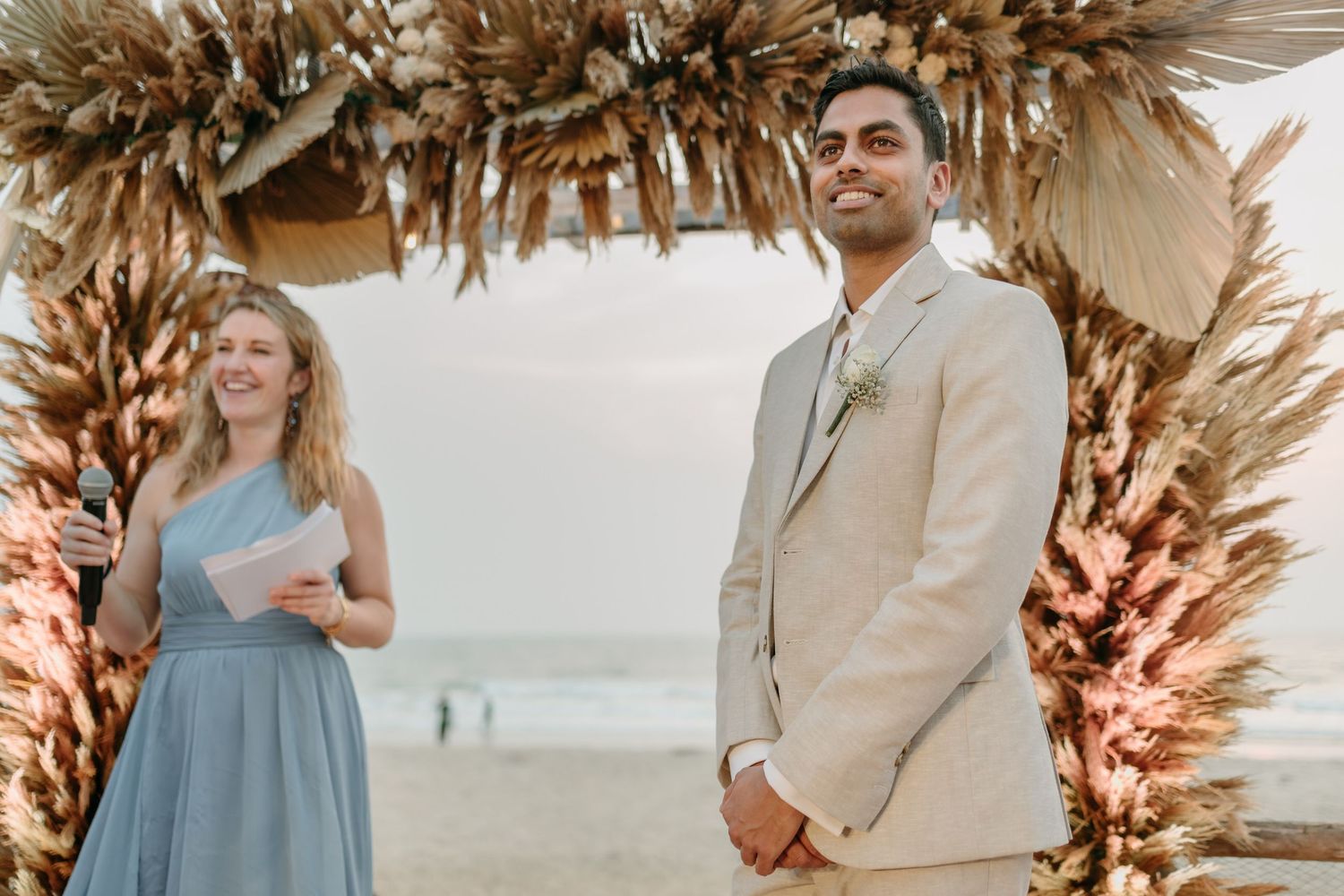 A couple stands under a dried palm arch at a beach wedding ceremony with soft tones and coastal backdrop.