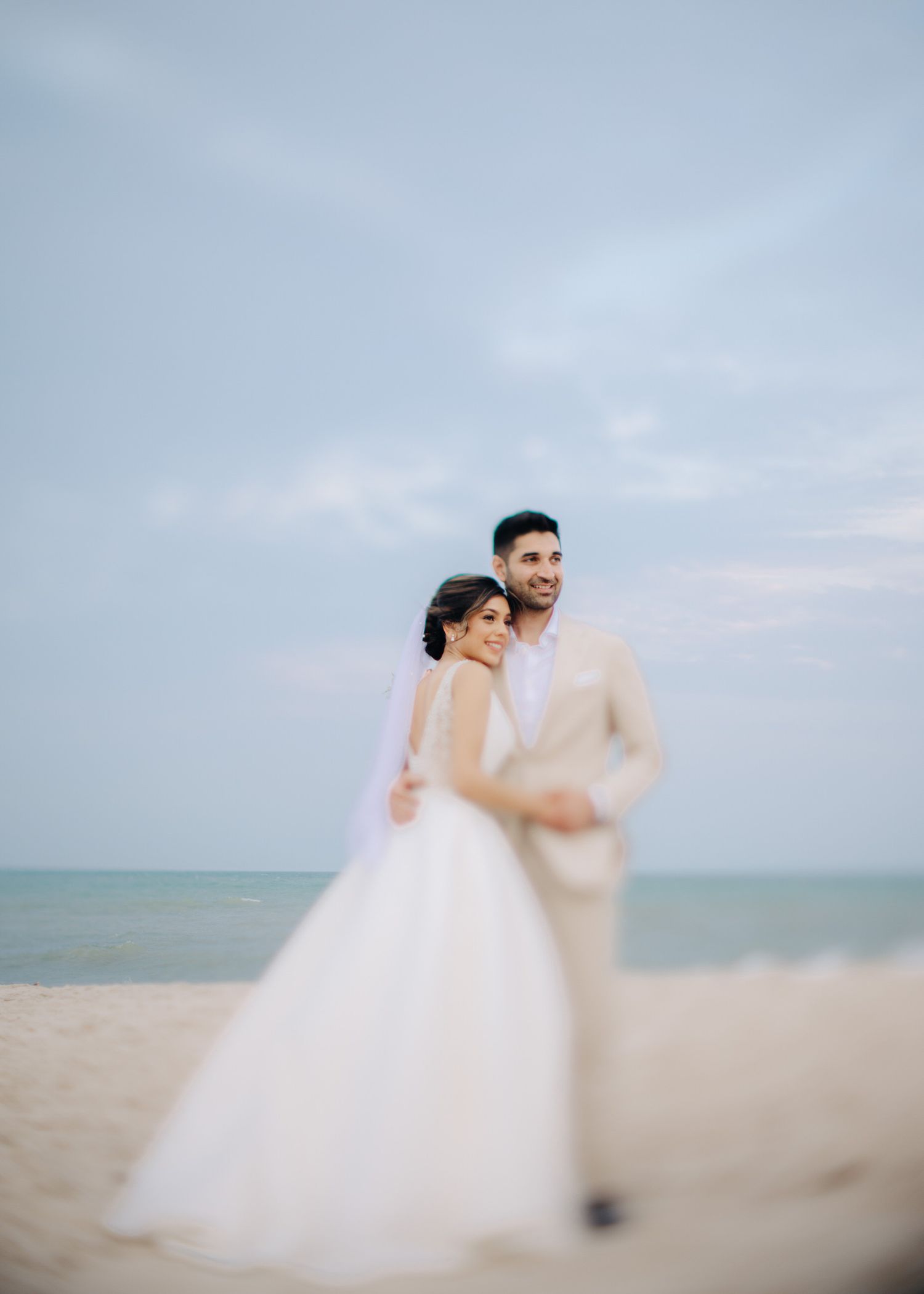 A couple in wedding attire embraces on a beach with soft cloudy skies in the background.