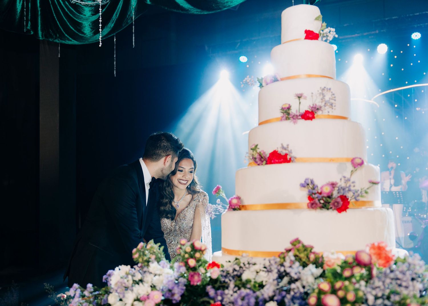 A multi-tiered white wedding cake decorated with red flowers displayed at a reception with blue stage lighting and floral decor.