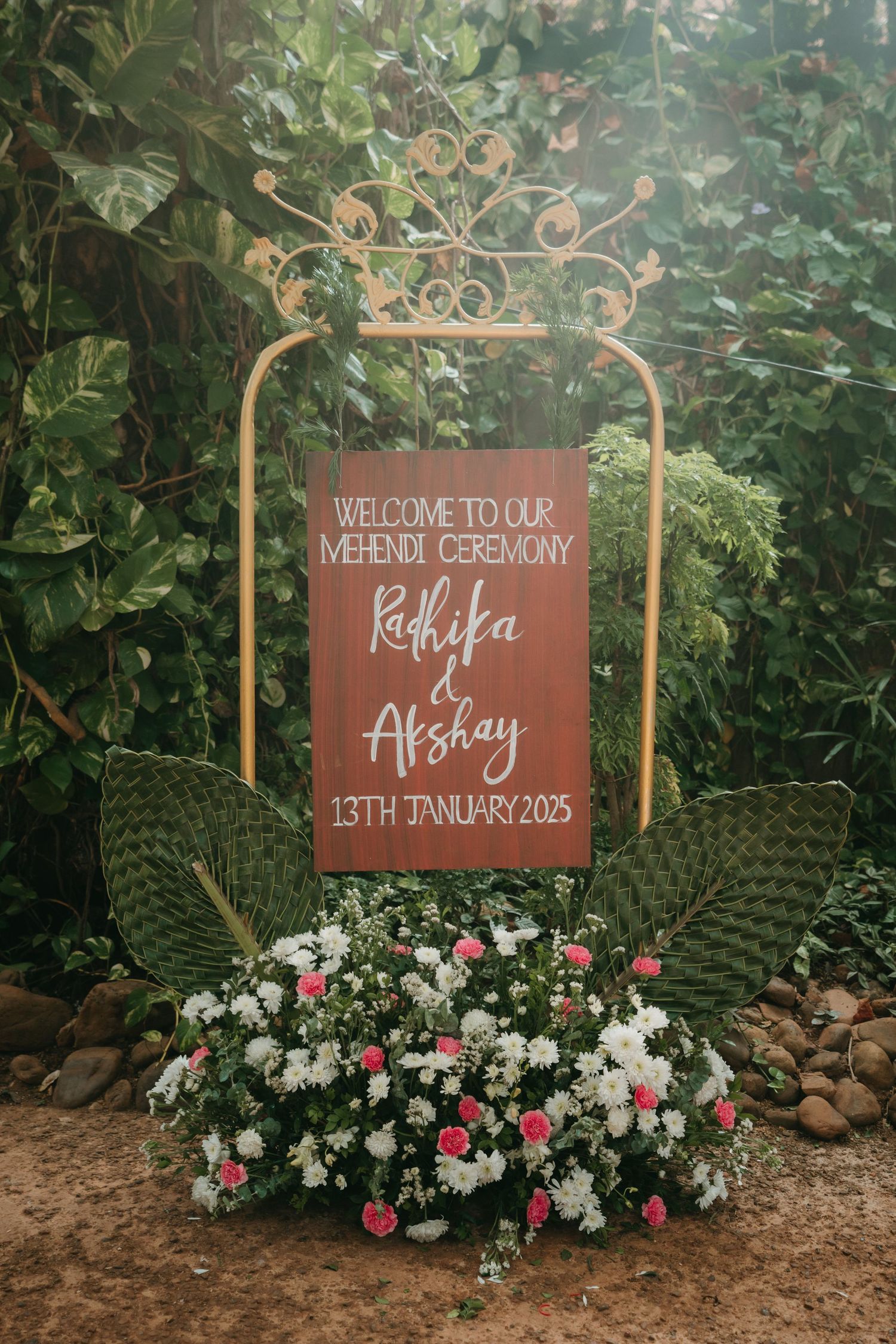 Elegant Mehendi ceremony welcome sign for Radhika & Akshay on January 13th, 2025, adorned with pink and white flowers.