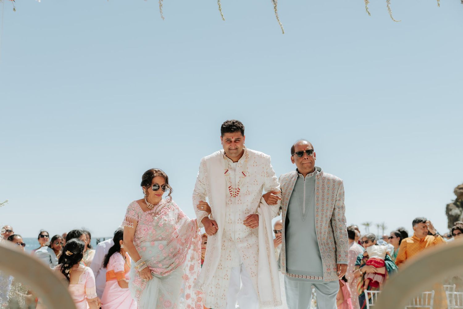 Groom in white sherwani walks down the aisle flanked by family at an outdoor Indian wedding ceremony.