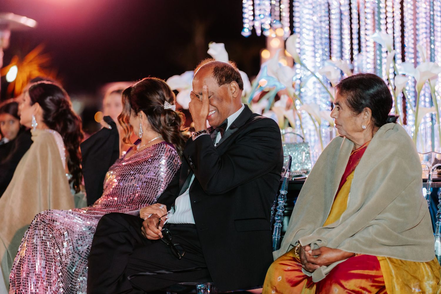 Emotional Indian wedding guests seated at outdoor reception, elder gentleman kissing bride's hand tenderly.