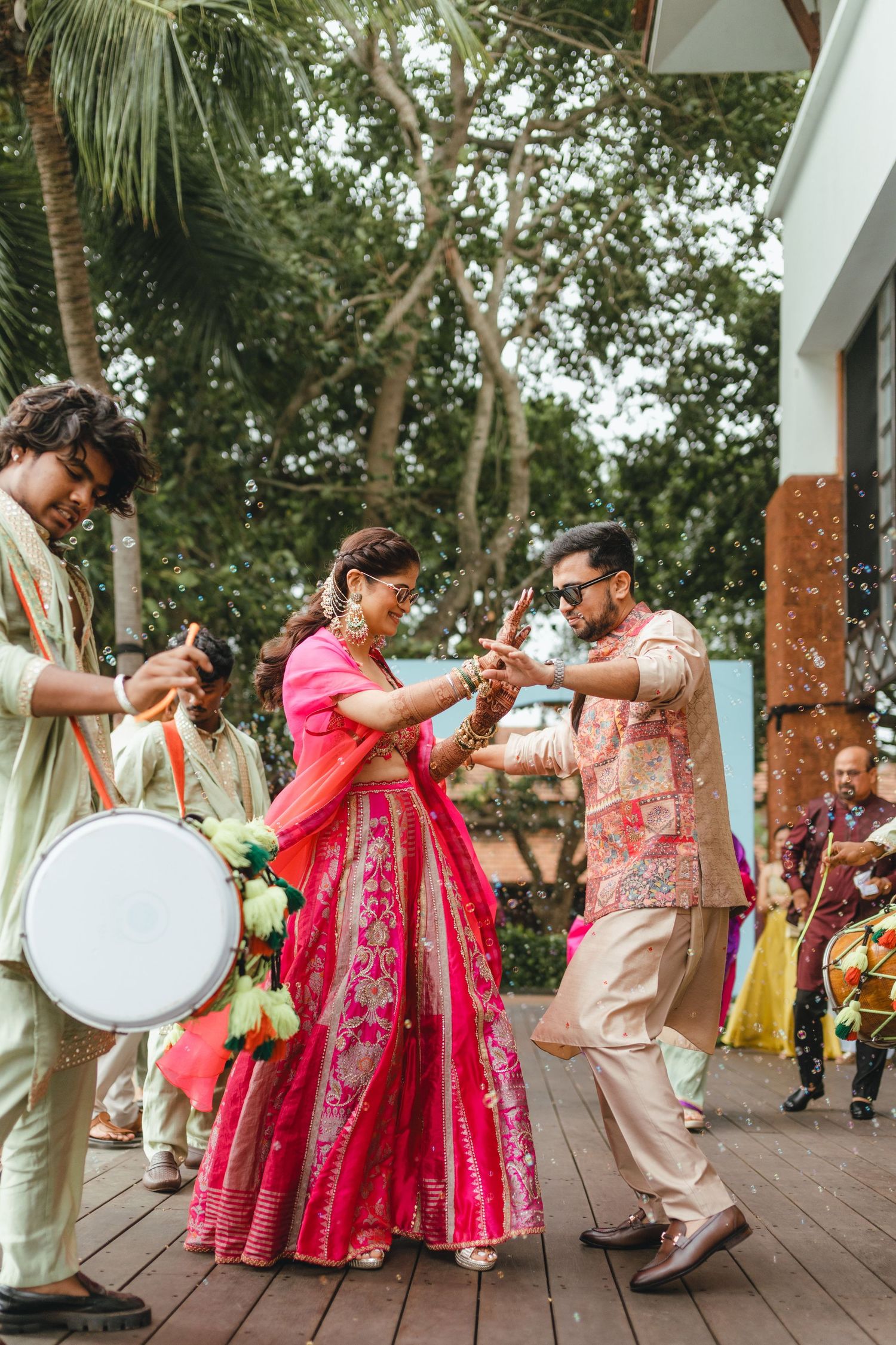 Bride in pink lehenga and groom in beige sherwani dance joyfully at Indian wedding celebration with dhol drummers.