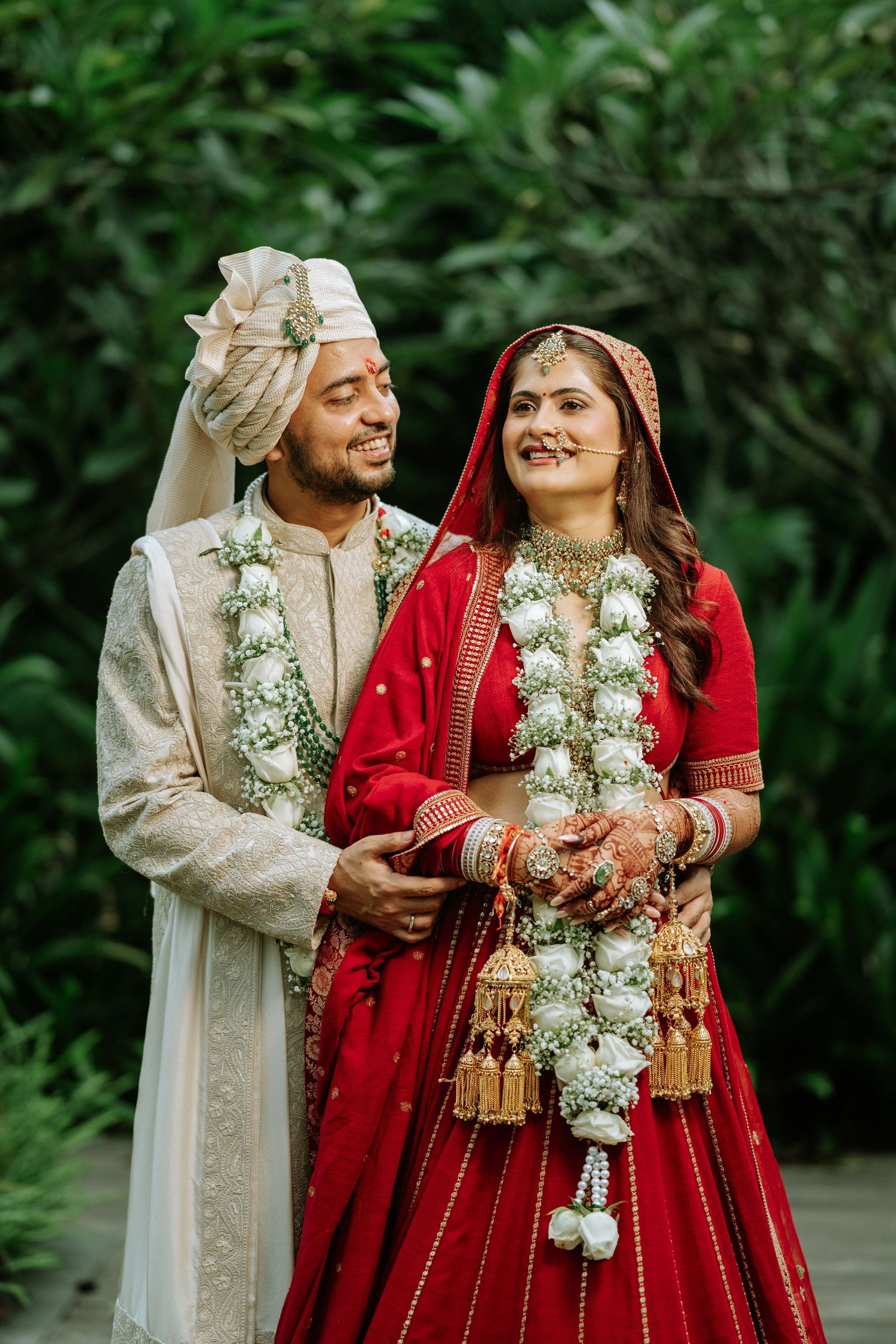Indian bride in red lehenga and groom in cream sherwani wearing floral garlands pose together at traditional wedding ceremony.