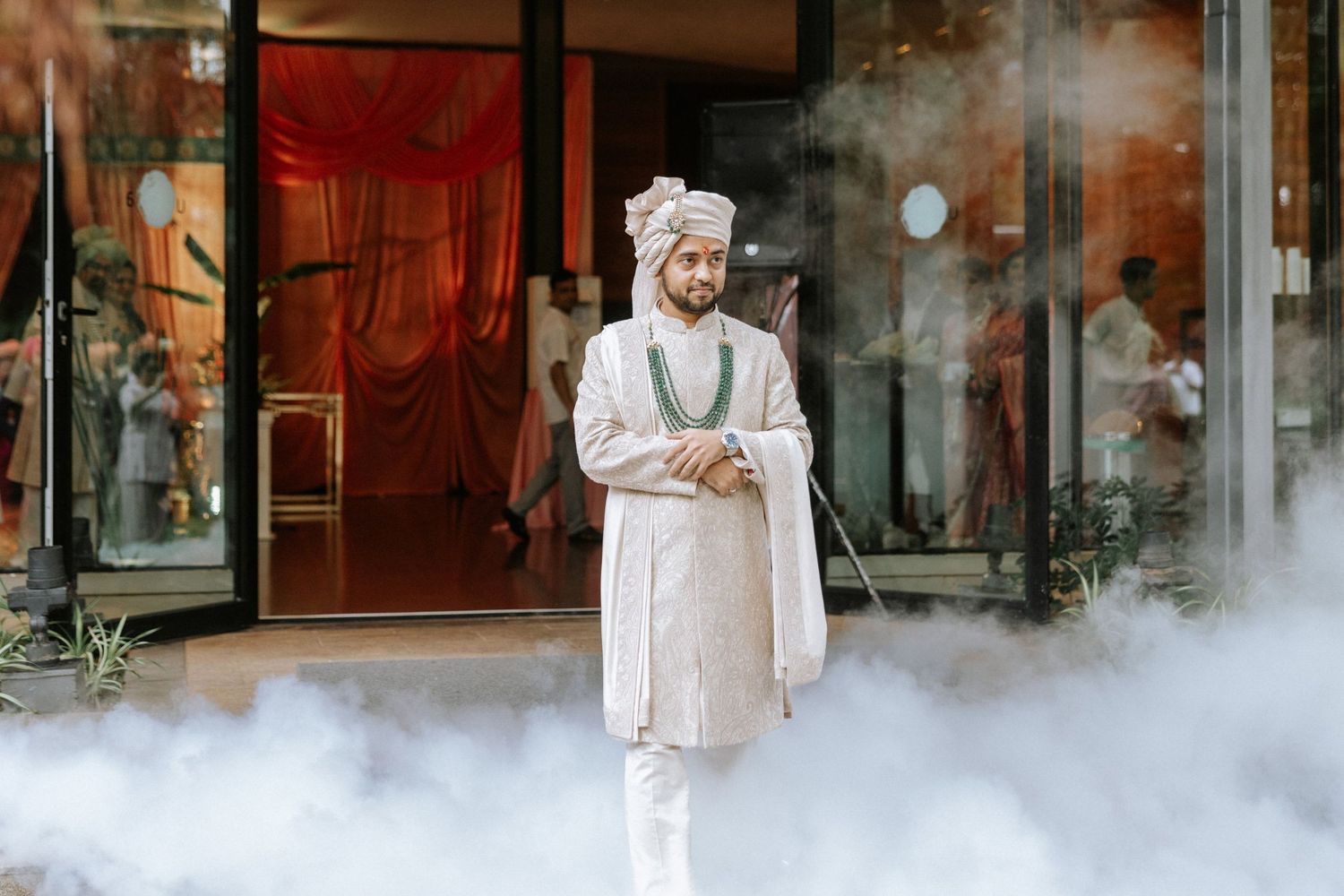Man in traditional Indian sherwani and turban stands confidently amid dramatic fog at an elegant venue.