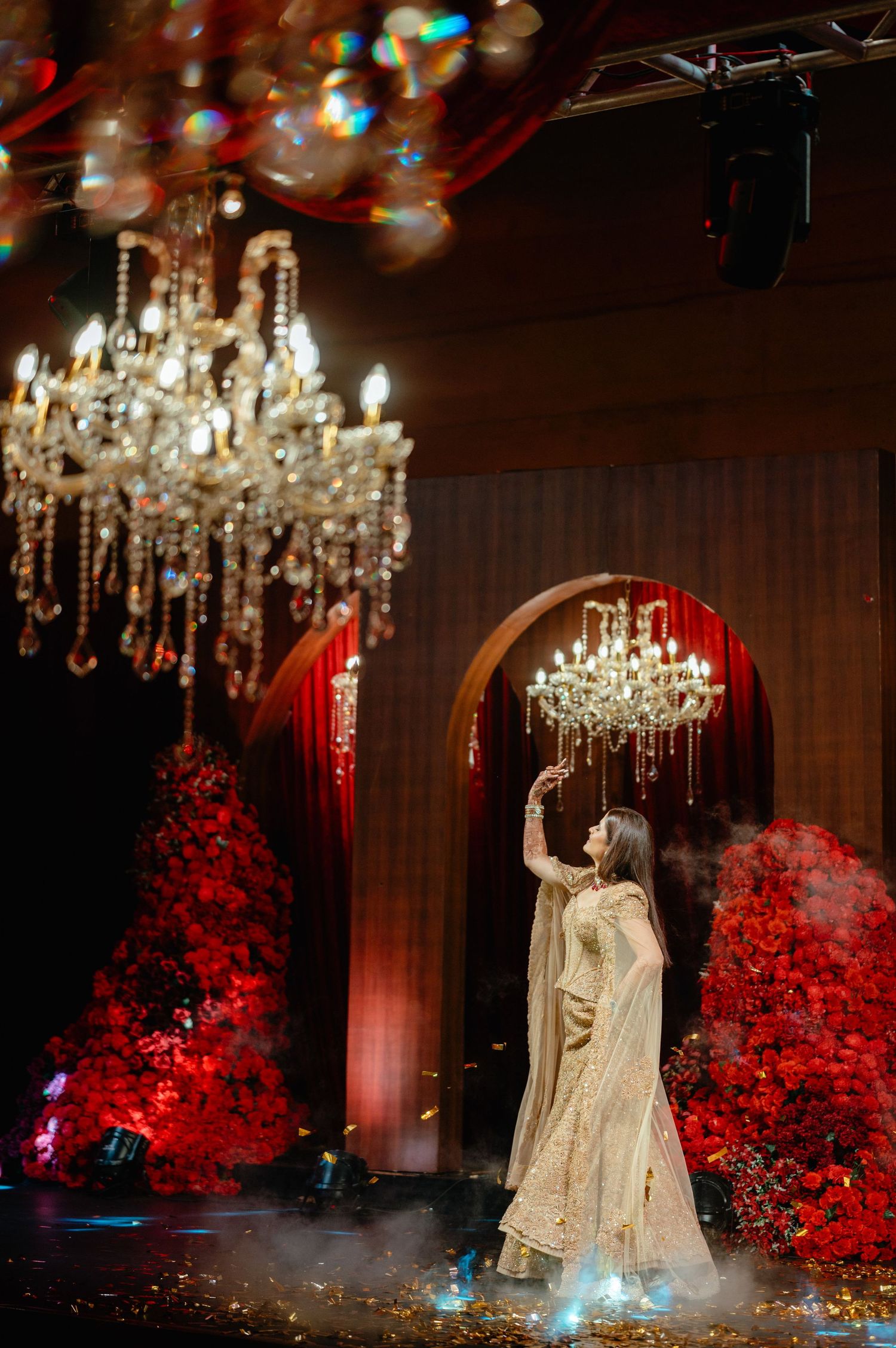 Bride in gold lehenga poses under crystal chandeliers amid red floral decor and dramatic stage lighting at wedding.