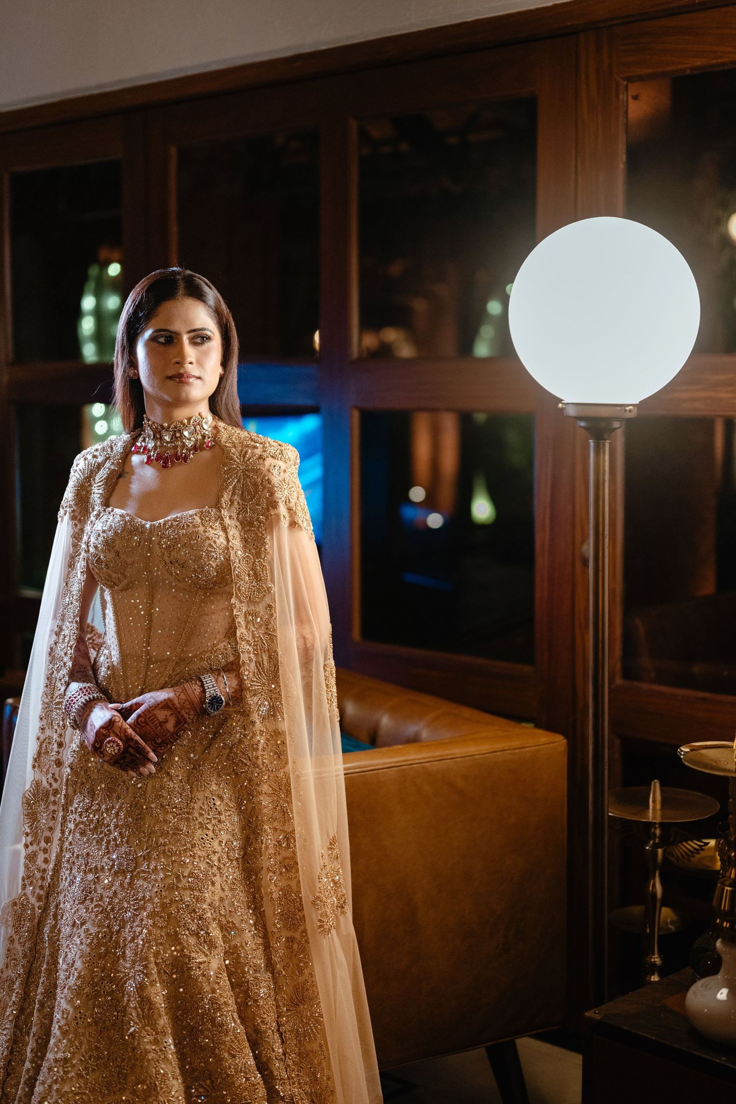 A bride in a golden embroidered lehenga with cape dupatta and red choker necklace poses elegantly indoors.