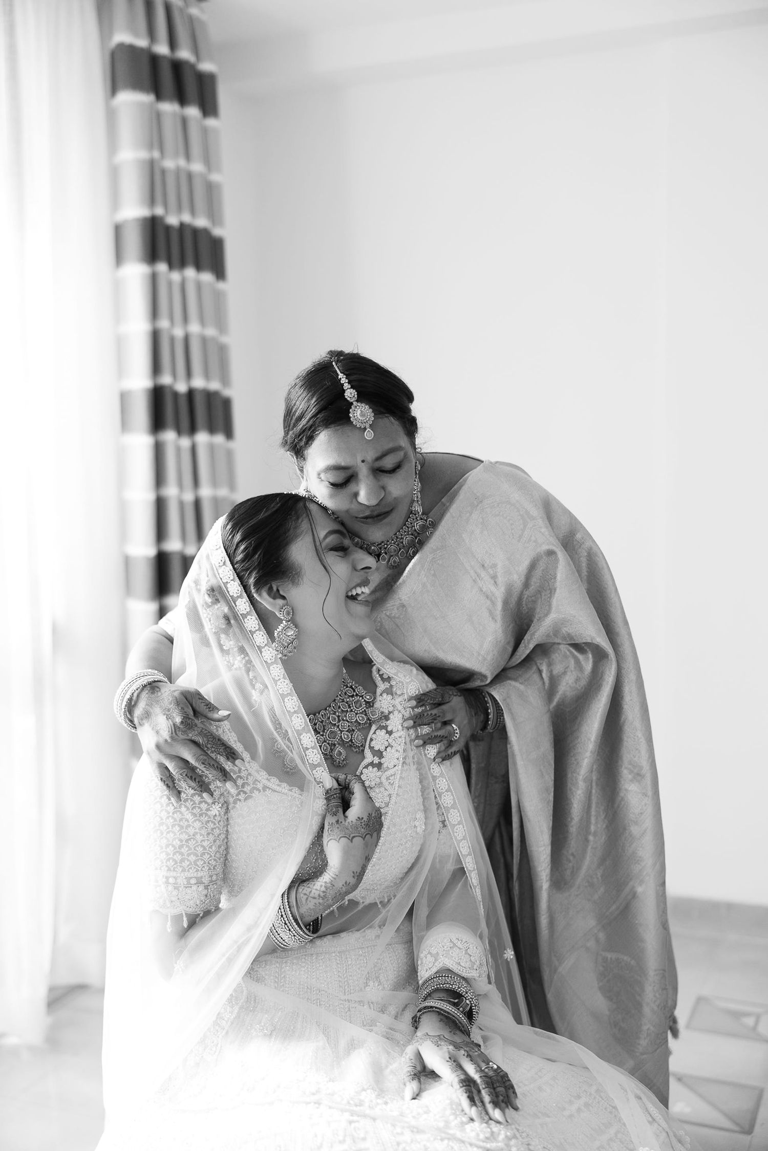 Black and white photo of a bride in traditional Indian attire being embraced and kissed by an older woman at a wedding.