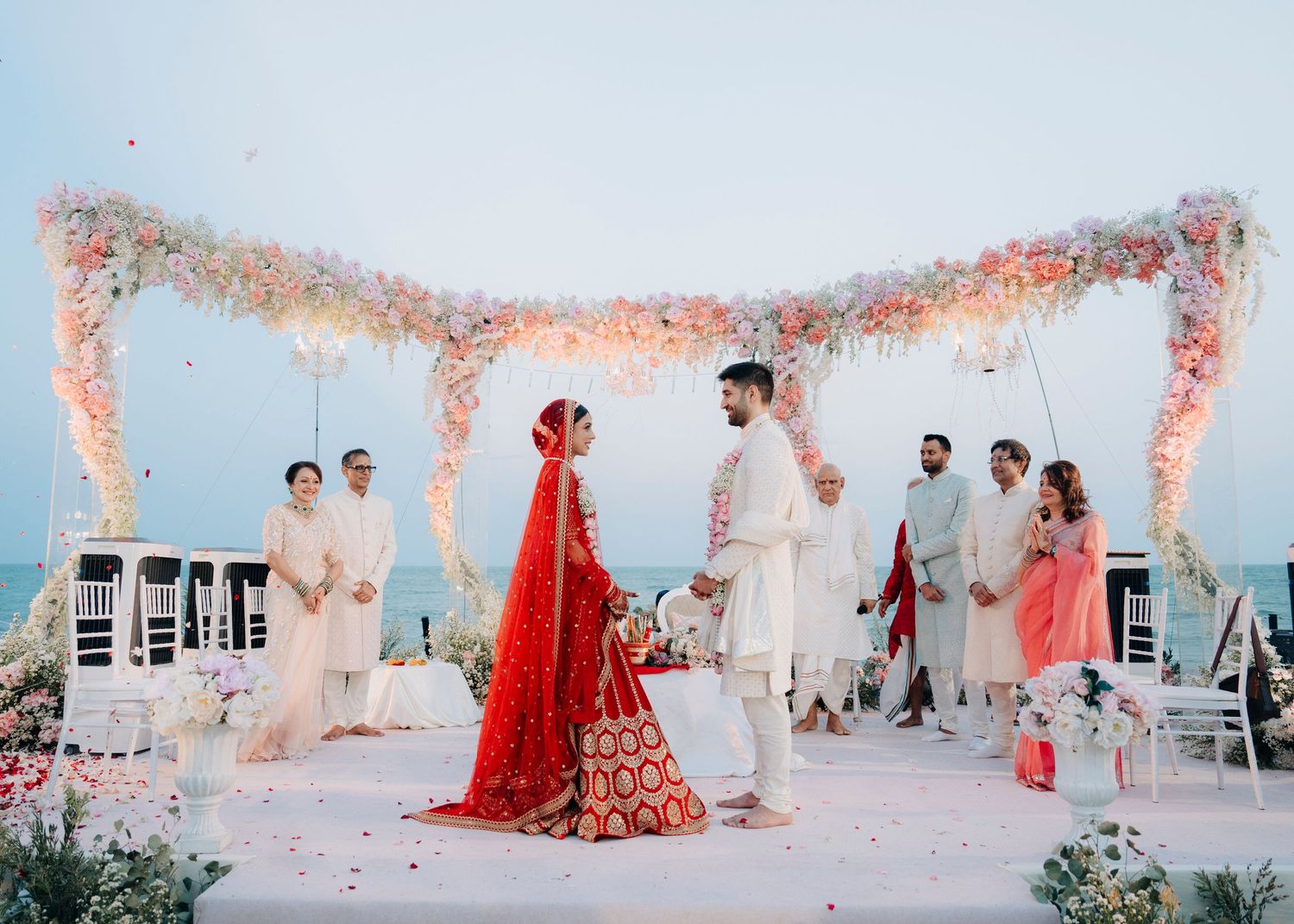 A stunning beachside wedding ceremony with floral decor and a couple in traditional red and white attire under a beautiful canopy.