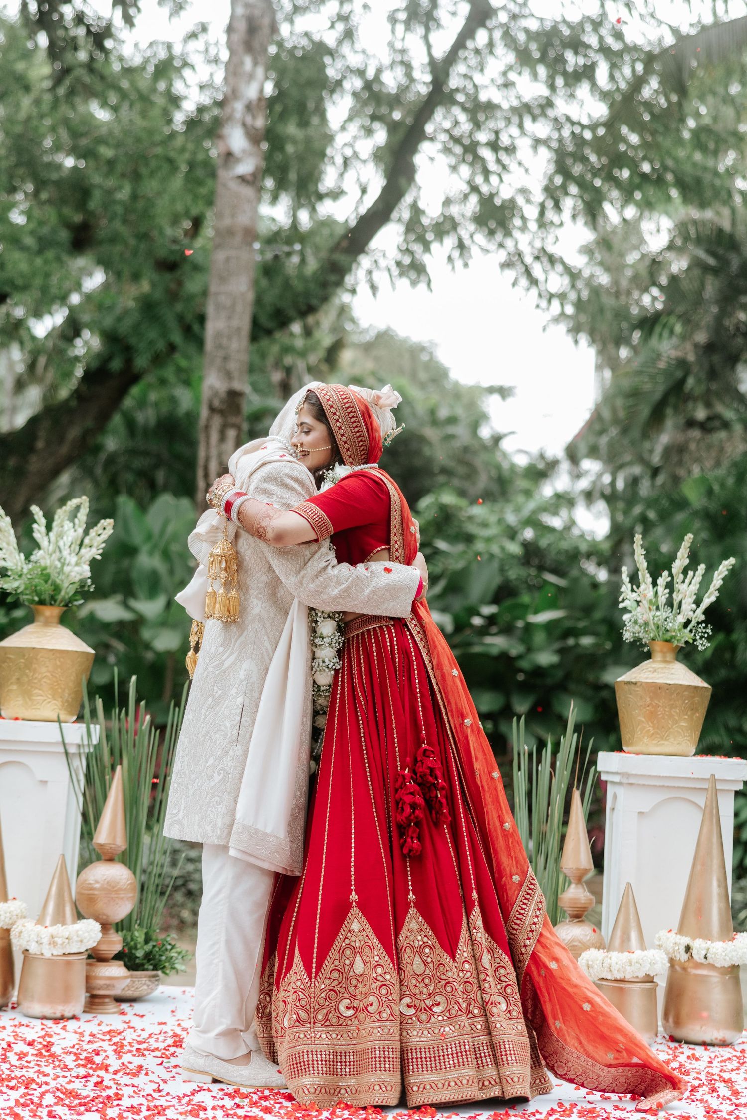 Indian bride in red lehenga and groom in white sherwani share a warm embrace at an outdoor wedding ceremony.