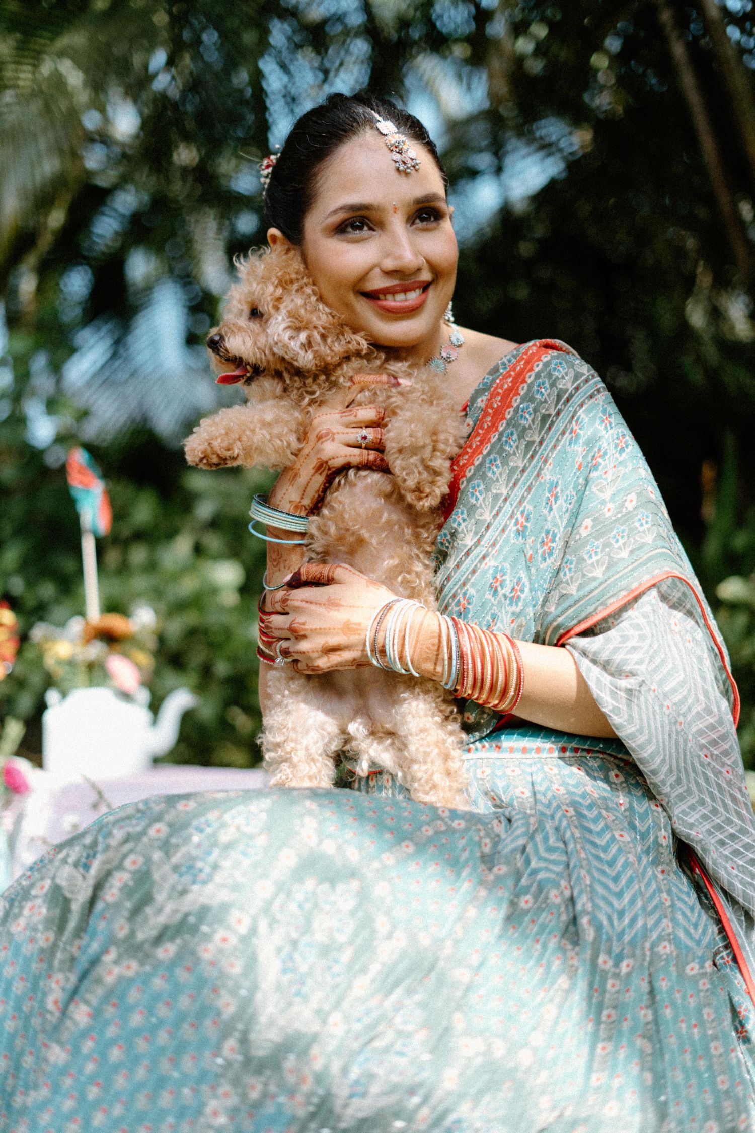 Smiling bride in light blue saree with bangles holds a fluffy brown poodle outdoors.