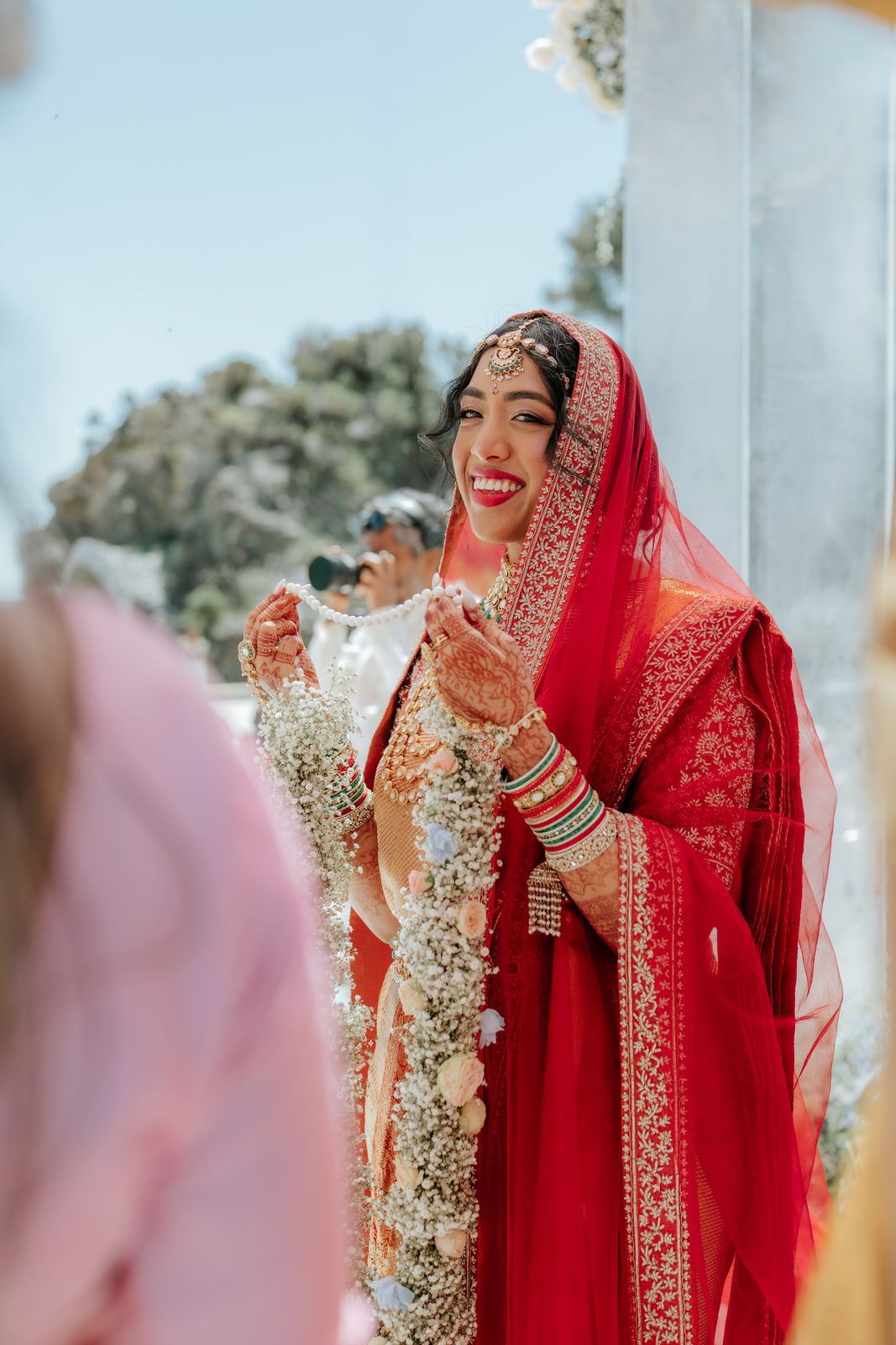 Smiling Indian bride in red lehenga holds flower garland during jaimala ceremony at outdoor Hindu wedding.