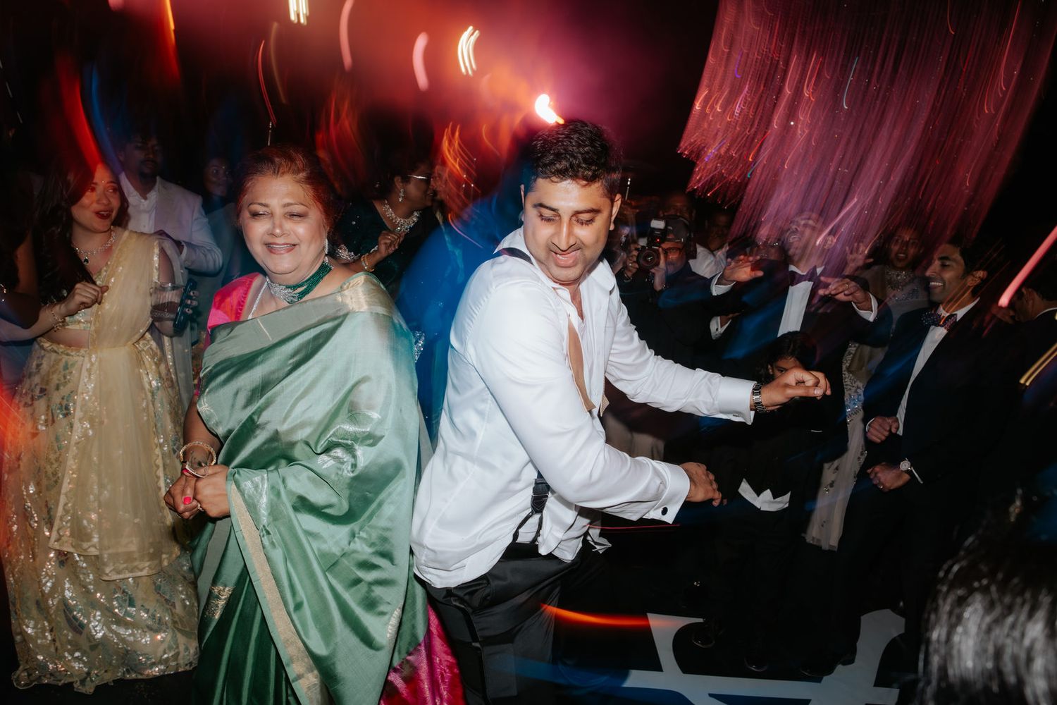 A woman in a green sari and a man in white shirt dance joyfully at a vibrant Indian wedding celebration.