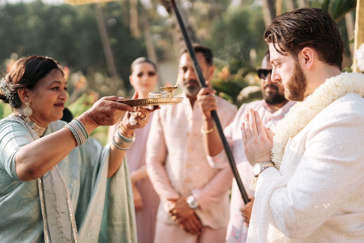 Indian wedding aarti ceremony with woman offering traditional ritual to groom wearing floral garland outdoors.