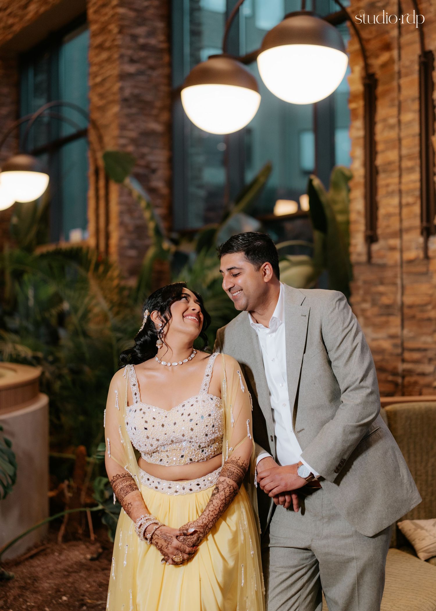 Couple in Indian wedding attire smiling together under pendant lights in a lush outdoor venue setting.