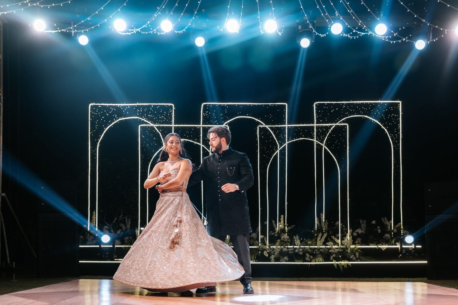 Couple sharing first dance on elegant stage with golden arch backdrop and dramatic blue stage lighting at wedding reception.