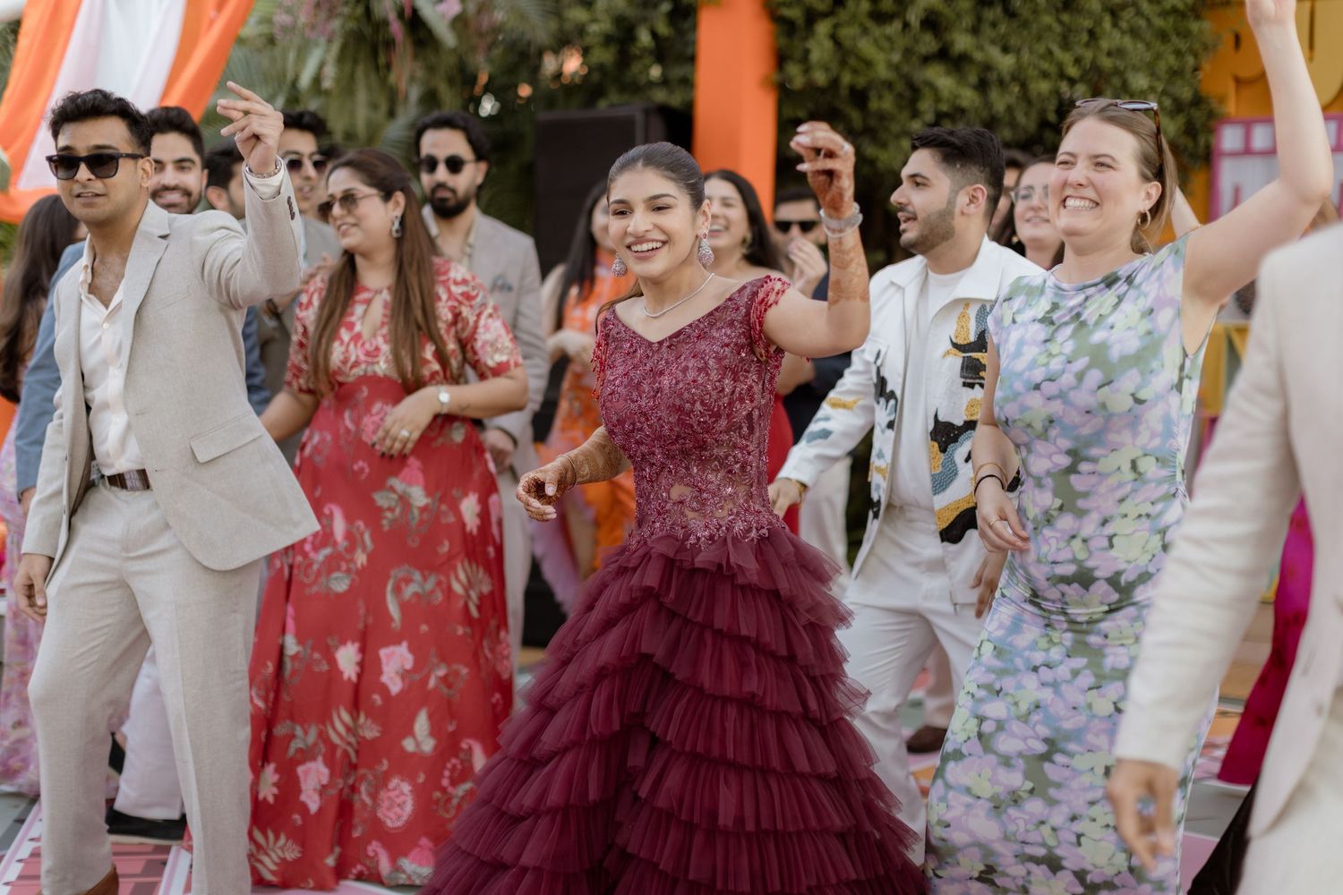 A group of wedding guests dance and celebrate at an outdoor reception with orange decorations and festive attire.