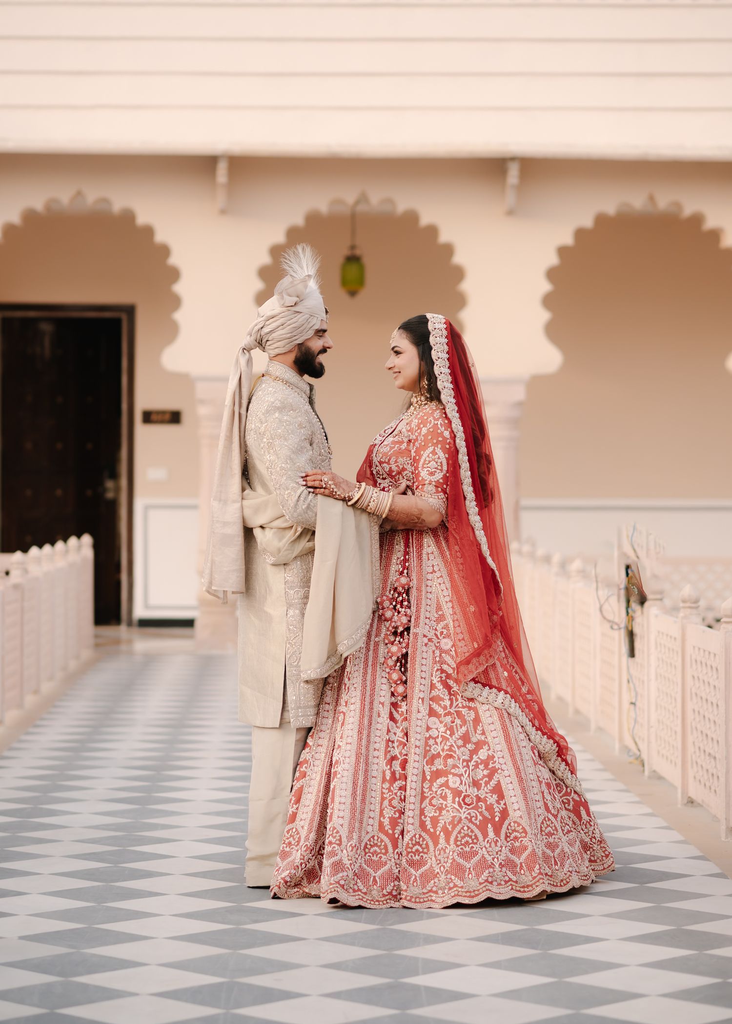 Indian bride and groom in traditional red lehenga and cream sherwani posing at ornate palace venue.
