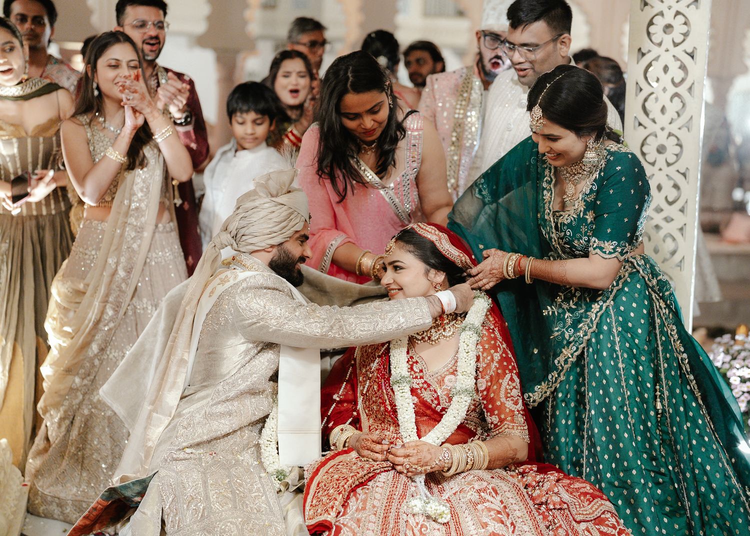 Bride in red lehenga receives sindoor during traditional Indian wedding ceremony surrounded by guests.
