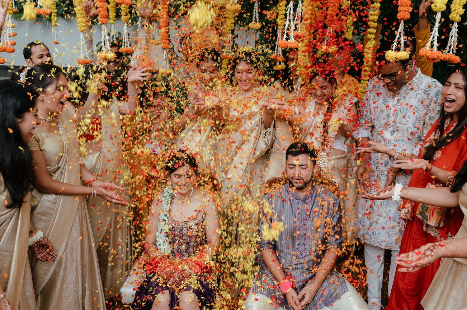 Colorful Indian wedding flower shower ceremony with bride and groom seated as guests toss vibrant petals and flowers.