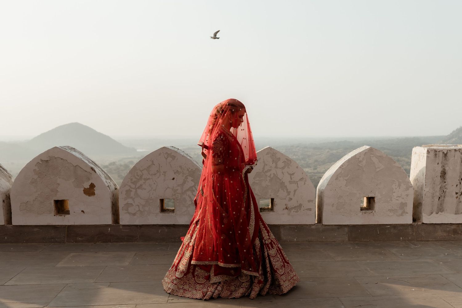 A bride in a red lehenga and dupatta stands on a fort terrace in Rajasthan, India, with scenic hills behind.